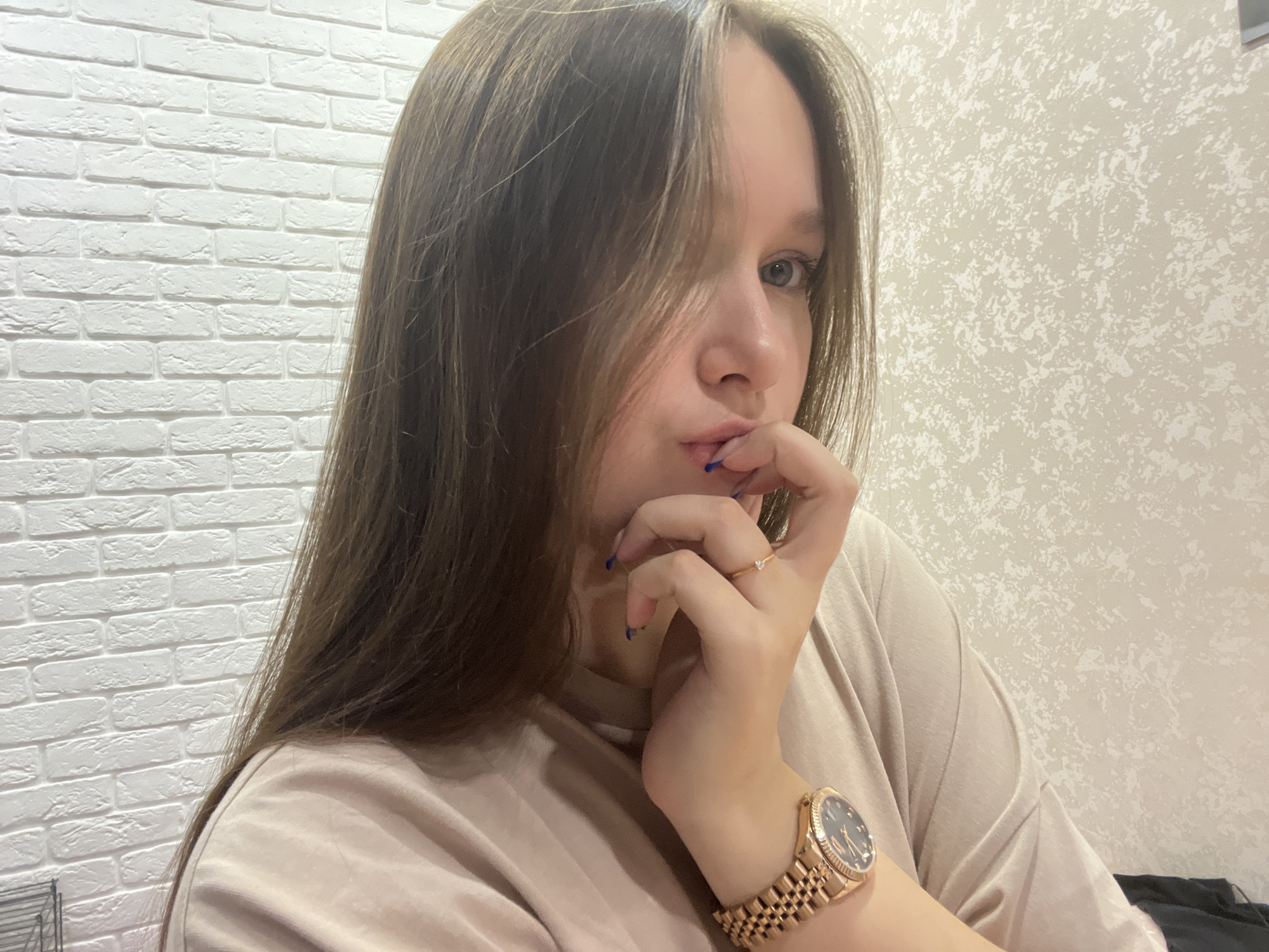 A young woman with shoulder-length brown hair poses for a selfie. Her hair is slightly disheveled, with strands falling across her face. She rests her chin on her hand, and her fingers are near her lips, which are painted a light pink. A gold ring adorns her ring finger. On her wrist, she wears a gold watch. She is wearing a light brown t-shirt.
The background consists of two walls. To the left is a white brick wall, and to the right, a wall with beige textured wallpaper.
There is no verifiable information to confirm that the image was taken in the city of Chaikovsky, Russia.