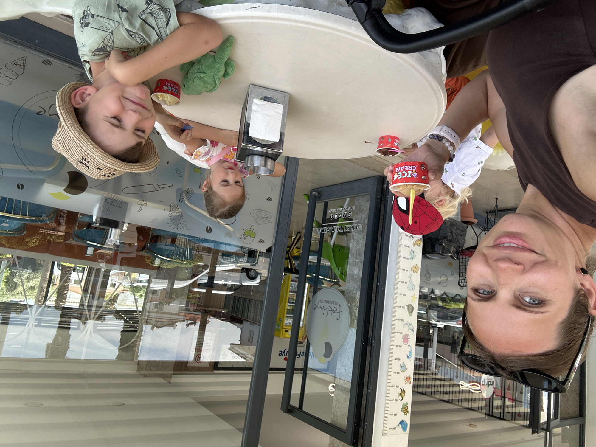 This is an outdoor scene in Fethiye, Turkey, possibly at the "Fethiye Dondurmacısı" (Fethiye Ice Cream Shop). An adult woman is taking a selfie at a table with two children. One child is wearing a hat and a green shirt with rockets and is holding a green plush toy. The other child is wearing a white shirt and an apron. All three are sitting at a round table with ice cream cups and a napkin holder. Behind them is the glass facade of the ice cream shop, reflecting the street scene outside. There is a height chart on the wall next to the shop entrance.
