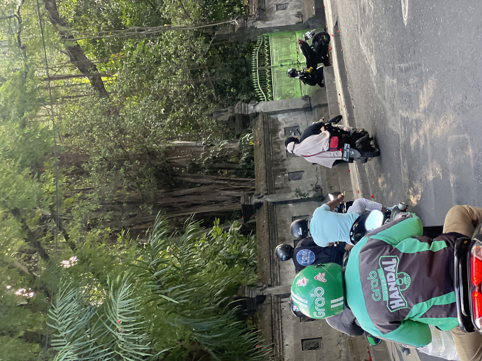 The image depicts a street scene in Kuta, Indonesia, likely in the late afternoon based on the lighting. The foreground shows several individuals on motorcycles, navigating a road. The rider in the immediate foreground wears a green helmet with "Grab" branding and a green and brown jacket also featuring "Grab" and "JUARA HANDAL" logos. Behind them, another rider sports a blue shirt and a black helmet. Further back, a motorcycle carries two passengers, one wearing a white headscarf and the other a dark helmet. A black motorcycle is parked on the side of the road to the right, near a green metal gate. The background is dominated by lush, dense foliage and the distinctive aerial roots of a large banyan tree, suggesting a tropical environment. A stone wall with ornate carvings runs along the side of the road, bordering the vegetation. The overall atmosphere is that of a busy urban street in a tropical locale.