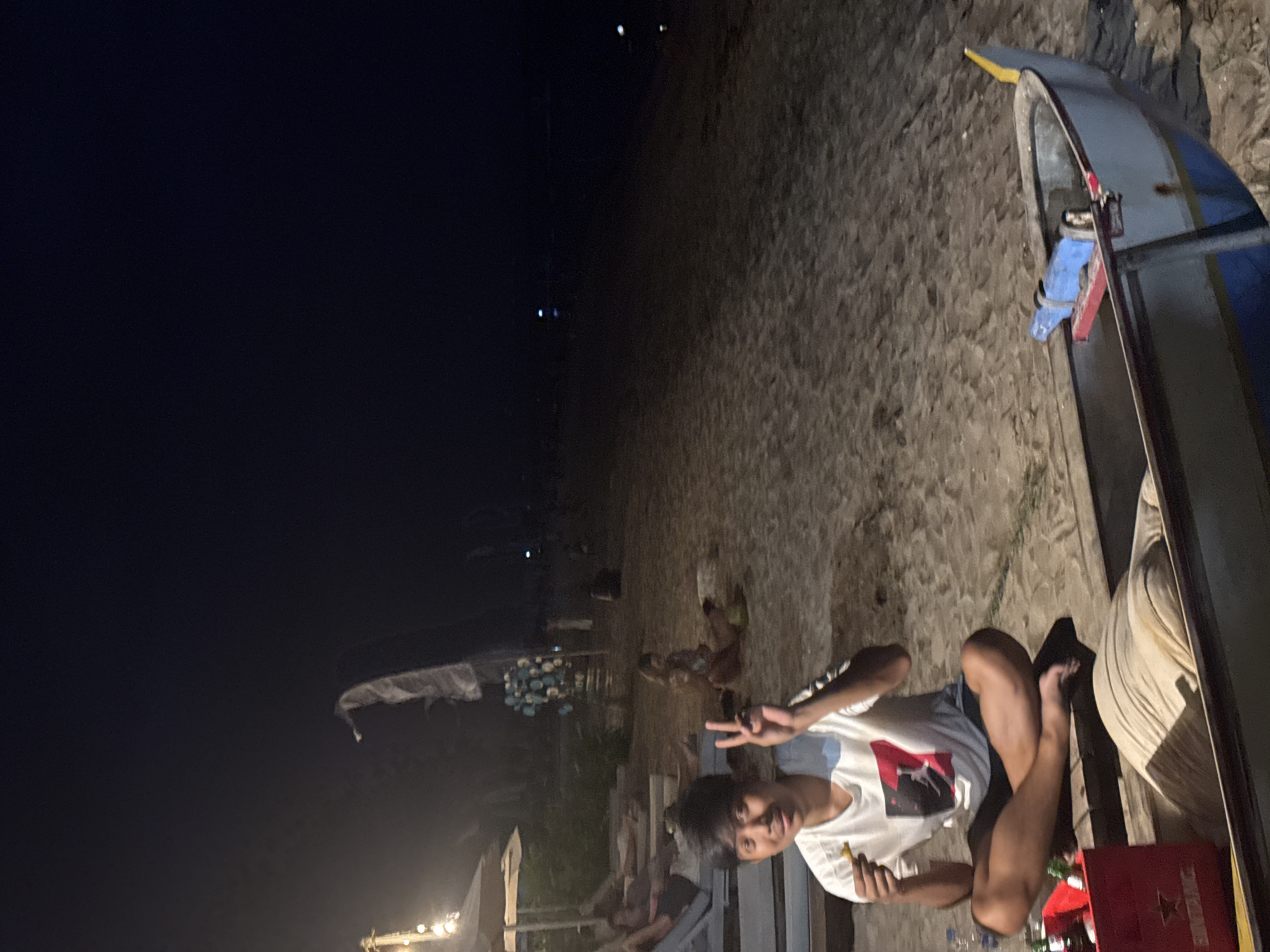 The image shows a beach scene at night, presumably in Denpasar, Indonesia. A young man is sitting in what appears to be a fishing boat on the sand, making a peace sign. He is wearing a white t-shirt and dark shorts. A red beer crate is visible nearby. In the background, the beach is lit by artificial lights, and several people and dogs can be seen. Fishing nets are visible in the distance. The sky is dark.