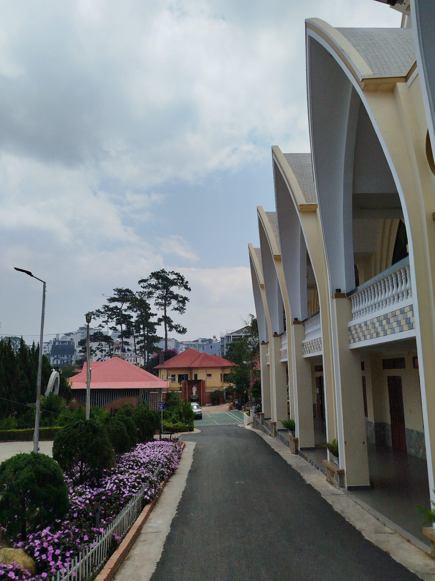 The image depicts a street in Da Lat, Vietnam. On the right side is a building with a series of arched supports, a tiled roof, and balconies. The street extends into the distance, lined with a flower bed, shrubs, and small trees on the left. In the background, there are buildings, trees, and a cloudy sky. The street is asphalt, and the lighting suggests it is daytime.