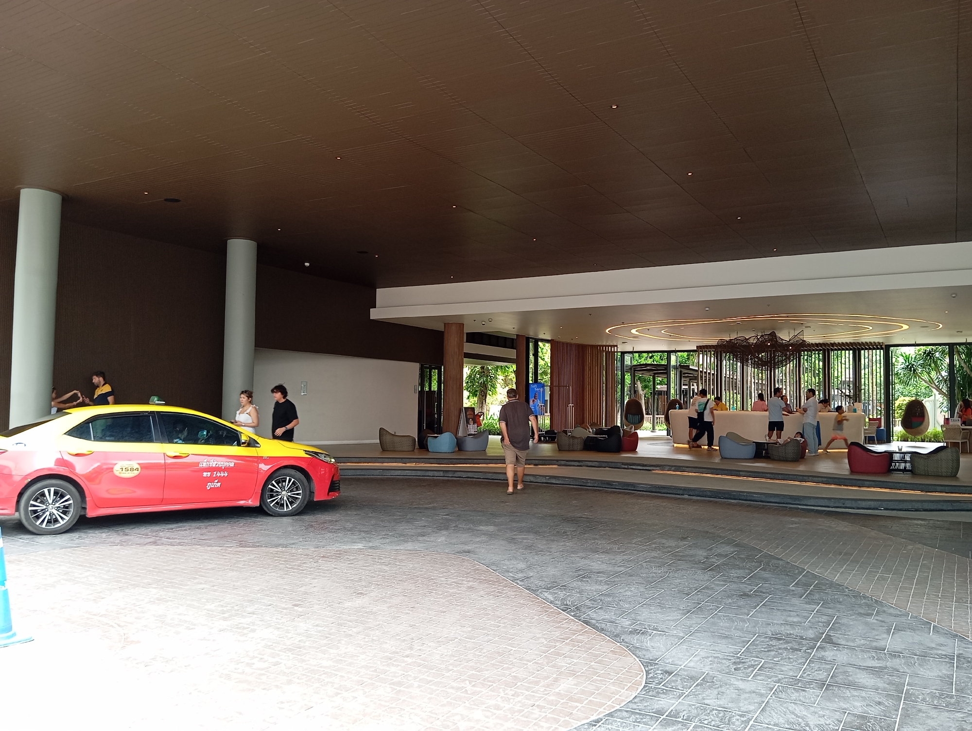 A red and yellow taxi is parked on a patterned driveway outside a building. The taxi has the number 1584 on its side. Two women and a man stand near the taxi. In the background, a man in a brown shirt and shorts walks away from the camera towards a building entrance. Inside the building, several people are visible at a reception desk and in a seating area with beanbag chairs and outdoor furniture. The building appears to be a hotel or resort lobby. The overall scene suggests a warm, tropical environment.
