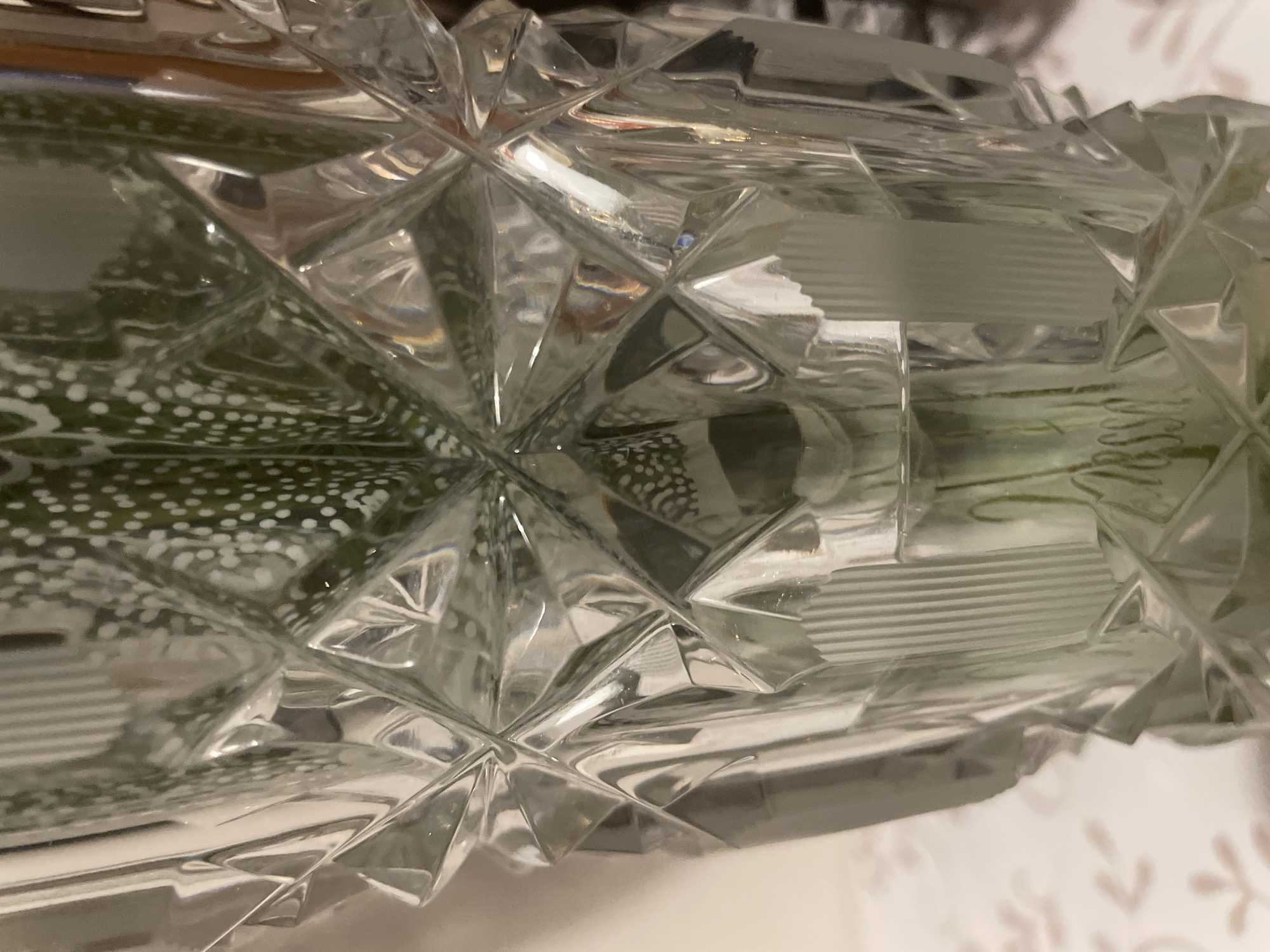 The image shows a close-up of a clear glass vase with diamond-shaped facets. Inside the vase, a plant with green leaves and white spots is visible. The plant appears to be a Haworthia, also known as a zebra plant. The vase is placed on a table with a patterned tablecloth. The background is blurred, but it appears to be an indoor setting. There are no people visible in the image. The image does not contain any information about the location in the city of Otradnoye, Russia.