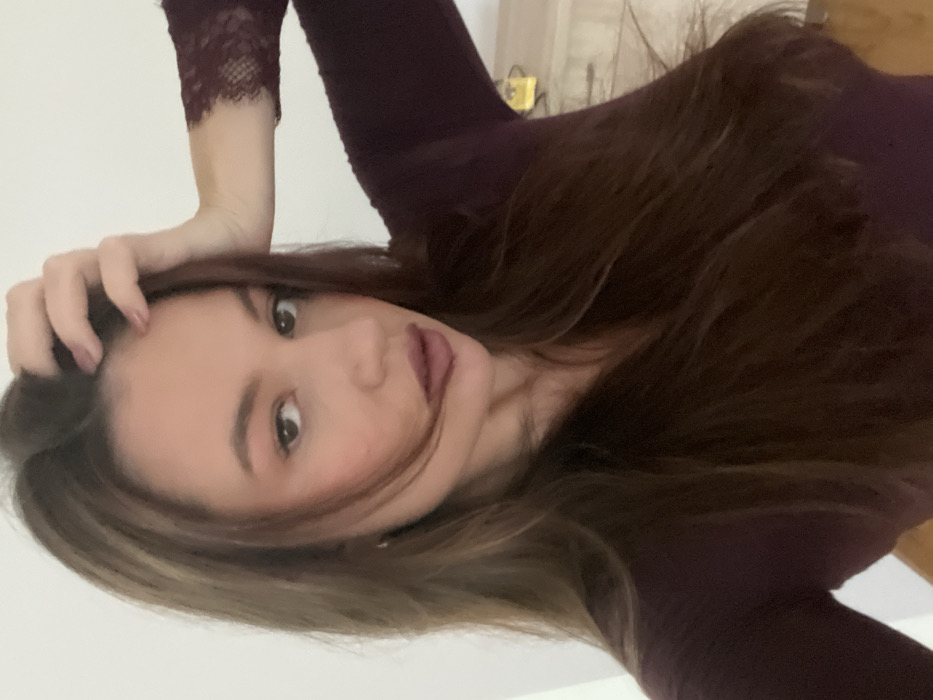 The image displays an adult female indoors, facing the camera. She possesses long, dark brown hair, parted, with some strands partially obscuring her face. Her lips are covered in a dark reddish-brown lipstick. She is attired in a fitted, long-sleeved top of a dark maroon hue; the visible portion of her right sleeve shows a lace cuff detail. Her left hand is raised, with fingers positioned on her hair above the forehead. The background comprises a plain white wall. To the subject's right, a partial view of a light-colored dresser or cabinet with visible drawer pulls is present, atop which rests a small, yellowish decorative object. A section of light-colored flooring is visible at the bottom right. The specific location within the city of Budva, Montenegro, is not discernible from the visual evidence.