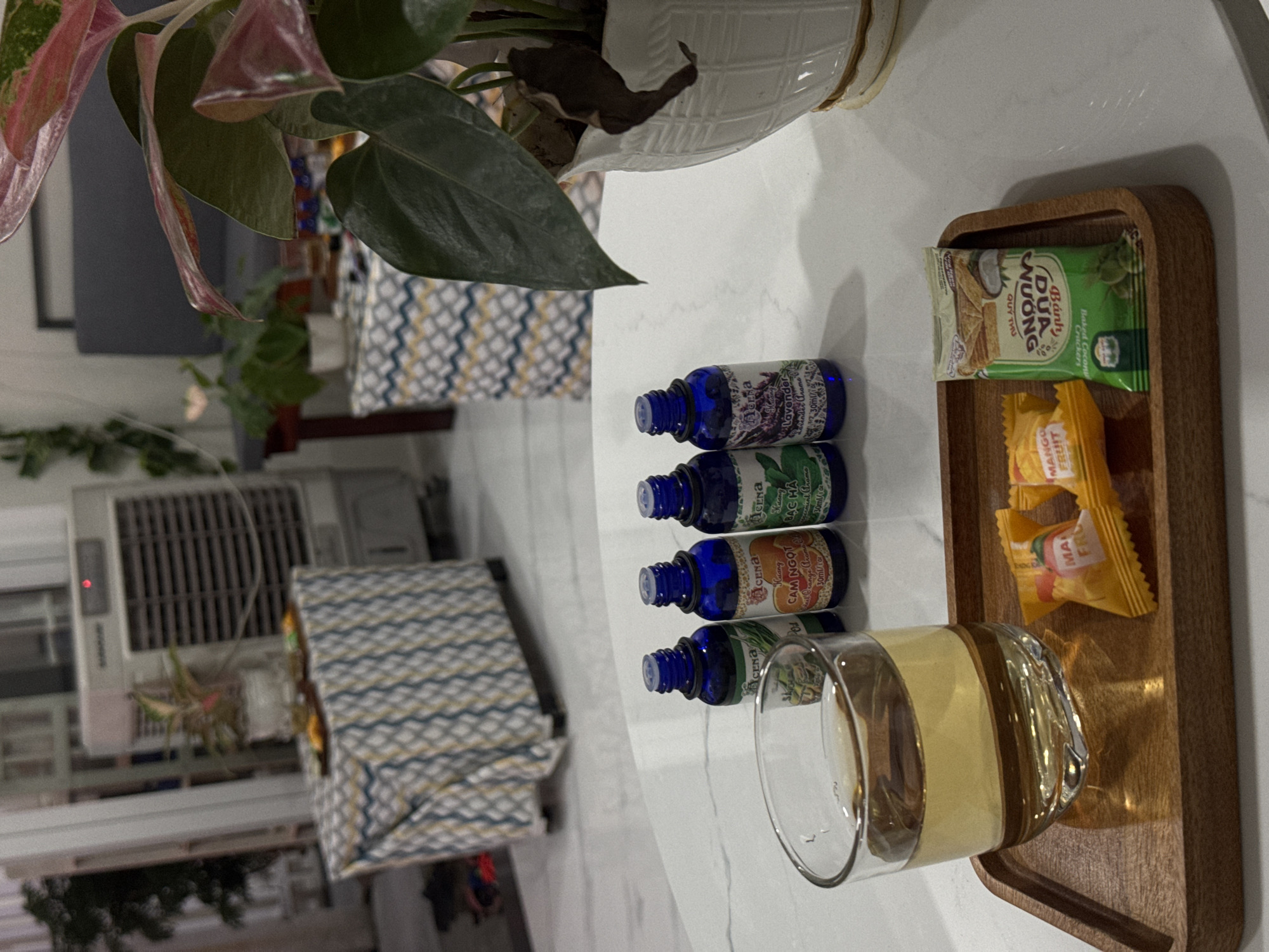 Here is a matter-of-fact description of the image:
The image shows a still life arrangement on a white surface, likely a table. The objects are arranged near an Anthurium plant in a white pot.
There's a wooden tray containing a partially filled glass of liquid (possibly tea or juice) and several snack packets. The snack packets include "DUA NUONG Banh Quy Thu" (Baked Coconut Crackers) and "MANGO FRUIT" candies.
Next to the tray are four small blue glass bottles with labels. The labels appear to read "ACENA Lavender," "ACENA Bac Ha," and "ACENA Cam Ngot," suggesting they contain essential oils or similar aromatherapy products.
In the background, there's a window air conditioning unit and a chair with a patterned cushion. The scene is indoors and appears to be well-lit.