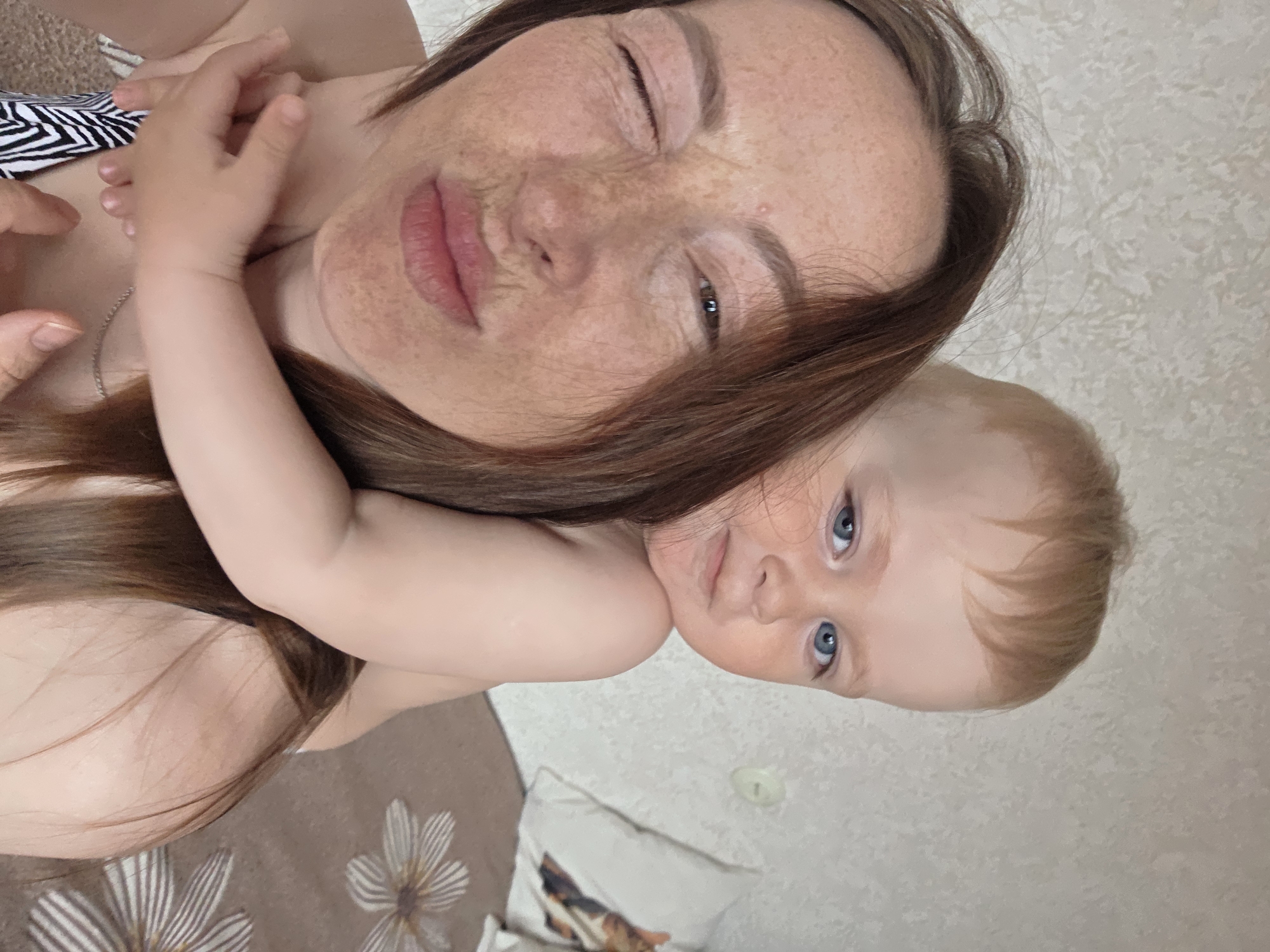 A woman and a baby are lying down. The woman is light-skinned with reddish-brown hair and freckles. Her left eye is closed. The baby is fair-skinned with blond hair and blue eyes, and is lying on the woman. They are on a bed with a beige bedspread and a white pillow with a floral pattern. The background is a beige wall. The location is Chaikovsky, Russia.
