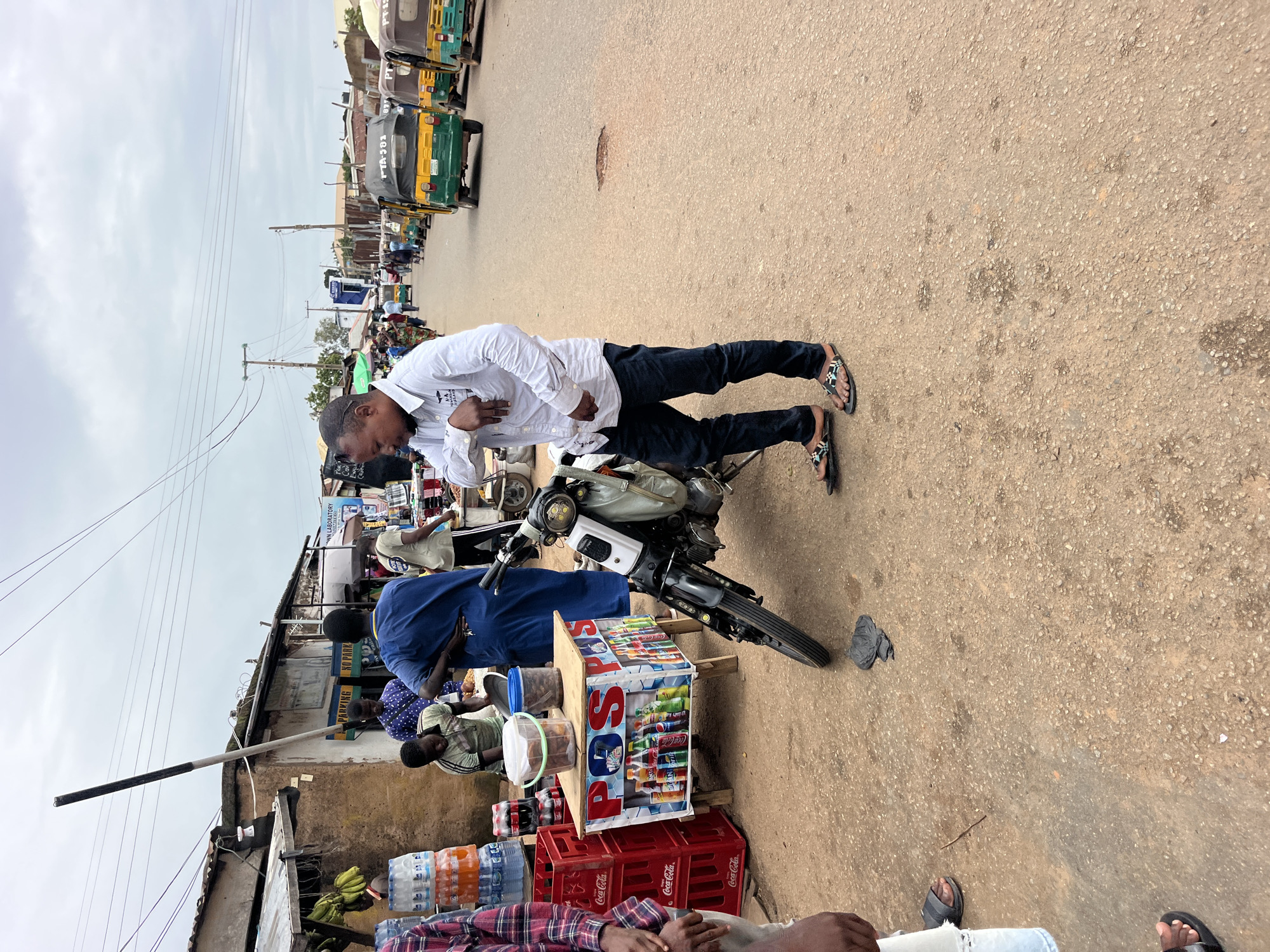 This is a vertically oriented, eye-level, medium shot of a street scene. A man in a white shirt and dark pants is standing with his left hand on his chest and his right hand resting on the handlebars of a motorcycle. The motorcycle is parked on a paved street. Behind the motorcycle, a market stall is set up, displaying various drinks in cans and bottles, as well as some plastic containers with food items. Red plastic crates labeled "Coca-Cola" are stacked beneath the stall.
In the background, several people are visible, some appearing to be vendors or customers. A line of colorful tricycles, commonly used as public transportation in some regions, are parked along the side of the street. The buildings in the background appear to be simple structures, some with corrugated metal roofs. Signs are visible on some of the buildings, with text including "WN LABORATORY," "PARKING," and "NO PARKING."
The sky is overcast, suggesting a cloudy day. The overall scene depicts a bustling outdoor market or street-side commercial area. The man in the foreground appears to be in a relaxed posture.
