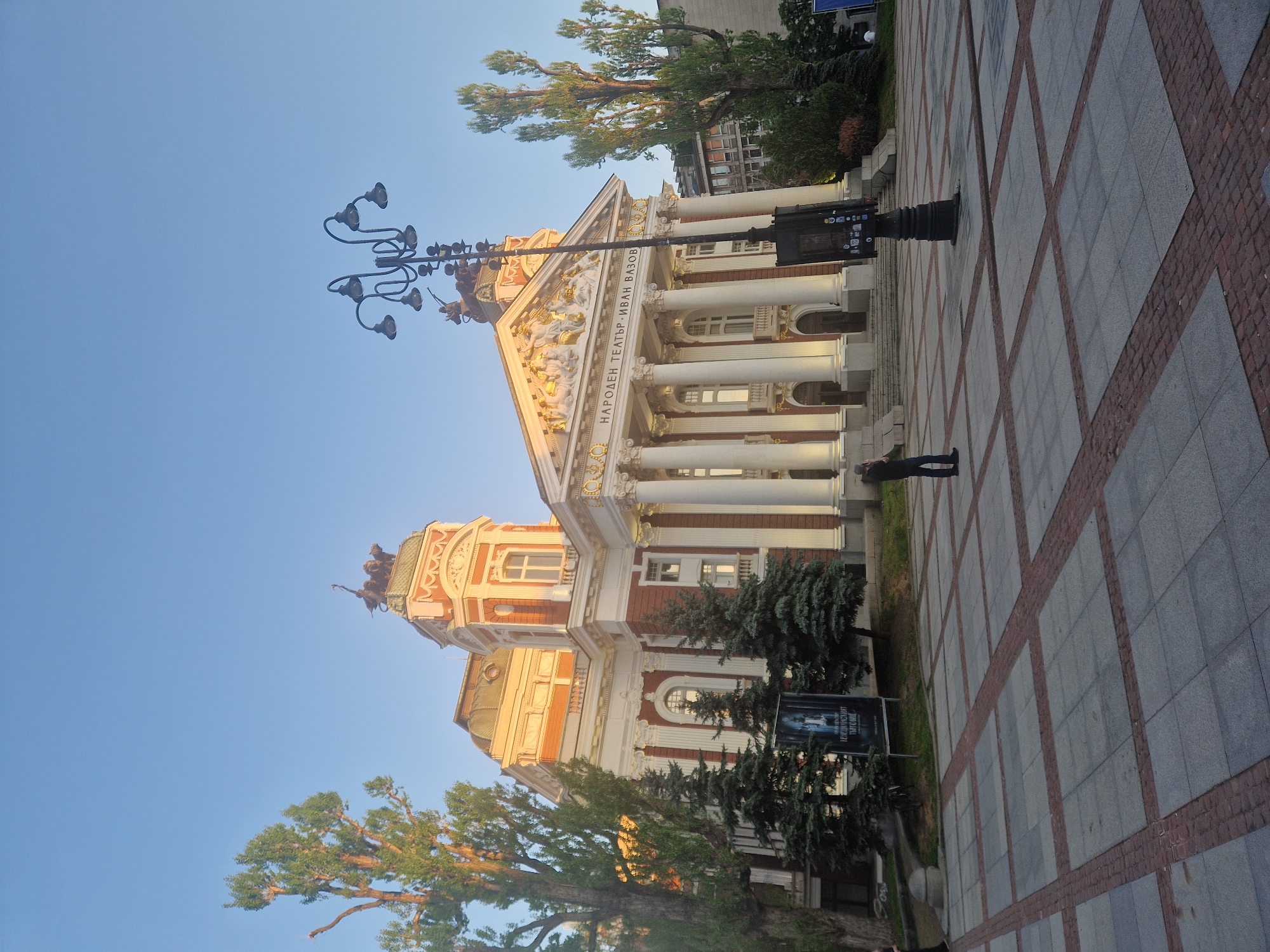 A low-angle, eye-level shot captures the National Theater "Ivan Vazov" in Sofia, Bulgaria. The building features a facade with white columns, reddish-orange accents, and ornate golden details. Text is inscribed above the columns, but the details are unclear. A man is standing on the paved plaza in front of the theater. To the left, a black ornamental street lamp with multiple curved arms and lamp heads is visible, along with the tops of green trees. A blue sky is in the background.