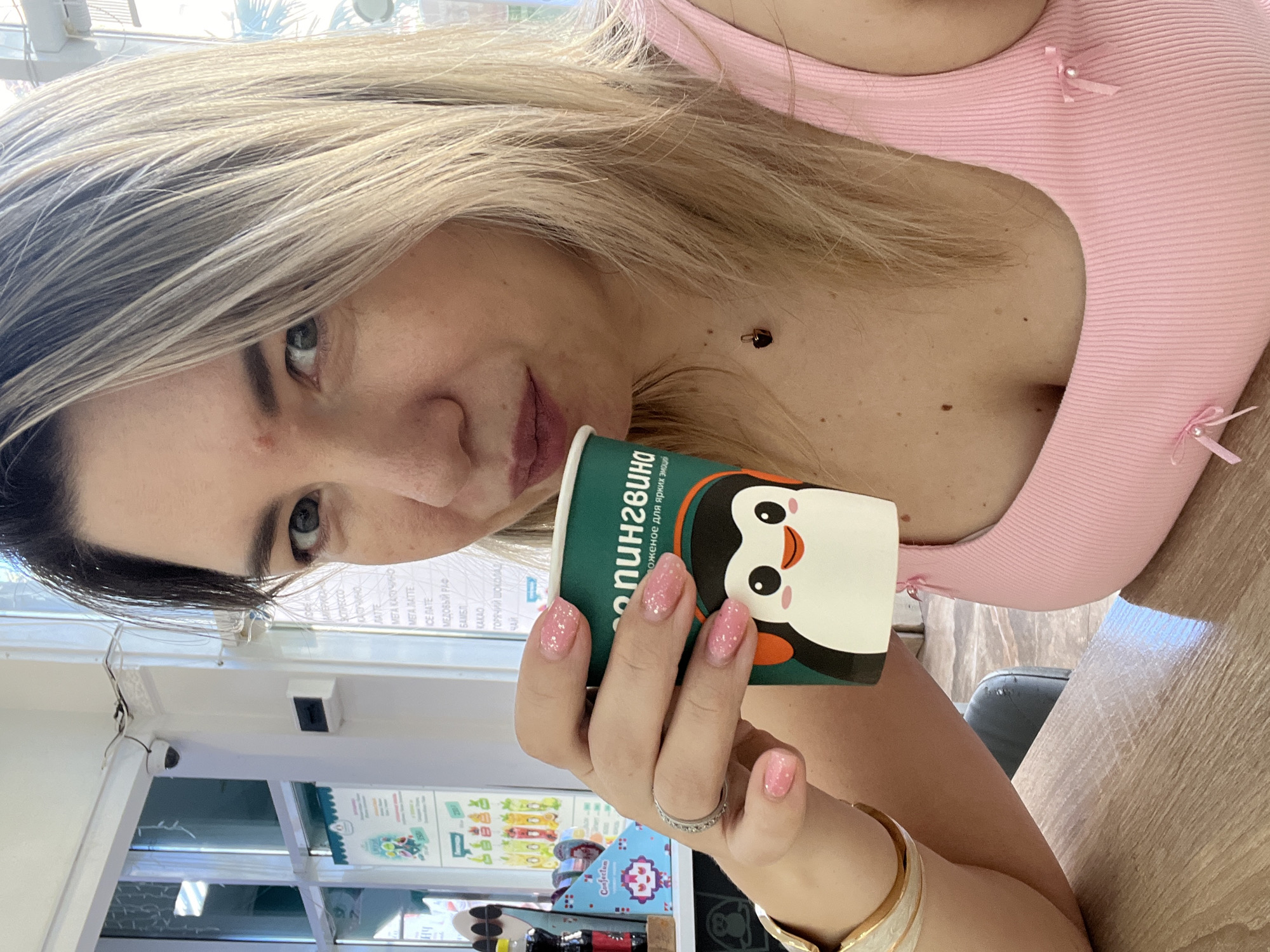 A woman with blonde hair and light skin is sitting at a wooden table, holding a green cup with a penguin design and drinking from it. She is wearing a pink tank top and a ring on her left hand. The background shows a bright room with windows, suggesting an indoor setting like a cafe or shop. The image appears to be taken in Sochi, Russia, as evidenced by the Russian text on the menu board in the background.