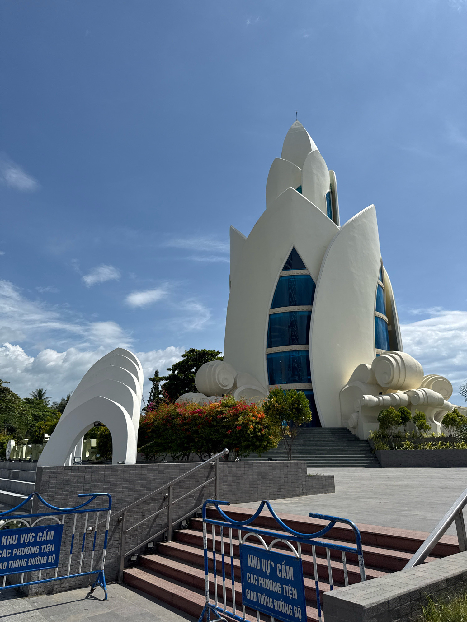 The image shows the exterior of the Long Son Pagoda in Nha Trang, Vietnam, on a sunny day with a blue sky and scattered clouds. The main structure is a large, white, lotus-shaped building with distinctive petal-like architectural elements and blue-tinted windows. To the left of the main building, there is a white, abstract sculpture that resembles several curved, overlapping arches. In the foreground, there are blue and white metal barriers with Vietnamese text, indicating a restricted area. Red-brown steps lead up to the pagoda, flanked by gray tiled walls and metal railings. Lush green trees and vibrant flowering bushes are visible around the structures. The overall scene depicts an architectural landmark in a well-maintained outdoor public space.