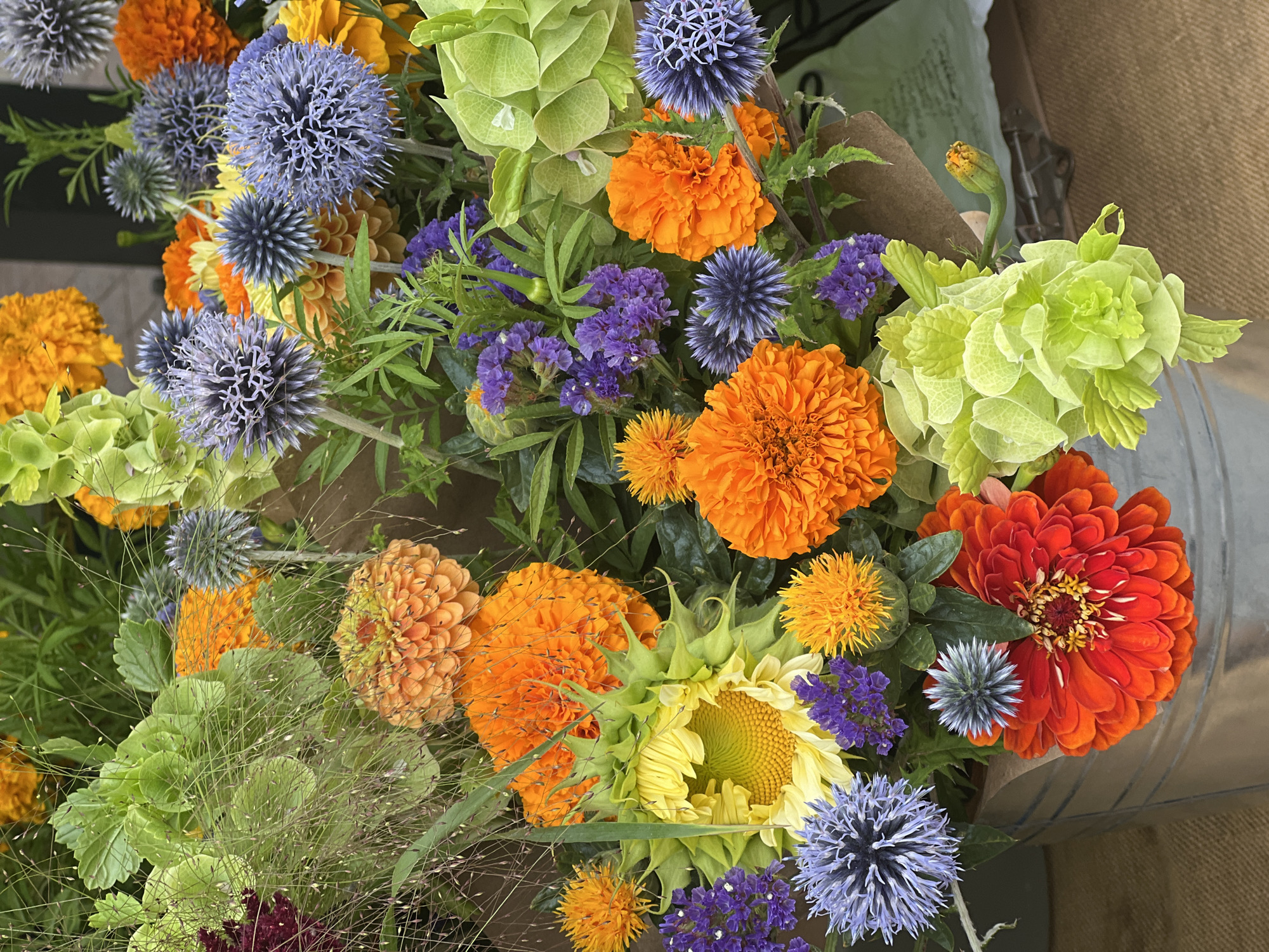 A detailed, close-up, vertical shot captures a vibrant bouquet of assorted flowers in a galvanized metal bucket. The arrangement is a riot of color and texture, featuring large, ruffled orange and red zinnia-like flowers, bright yellow sunflowers with green buds, spiky globe-shaped blue thistles, delicate purple statice, and fluffy, pale green ornamental kale. Interspersed throughout are wispy, thin stems of grass. The bucket itself is partially visible at the bottom, made of silver metal with a rustic, slightly dented appearance. The background is blurred, with hints of brown paper wrapping the flowers and a dark, indistinct area behind. The scene is shot outdoors, likely on a bright day, given the rich saturation of colors. There are no people visible in the image. The flowers appear to be part of a market stall or shop display, but no specific signage or identifying marks are present to confirm the location. Without further information or visible landmarks, the exact location in Revelstoke, Canada cannot be determined.