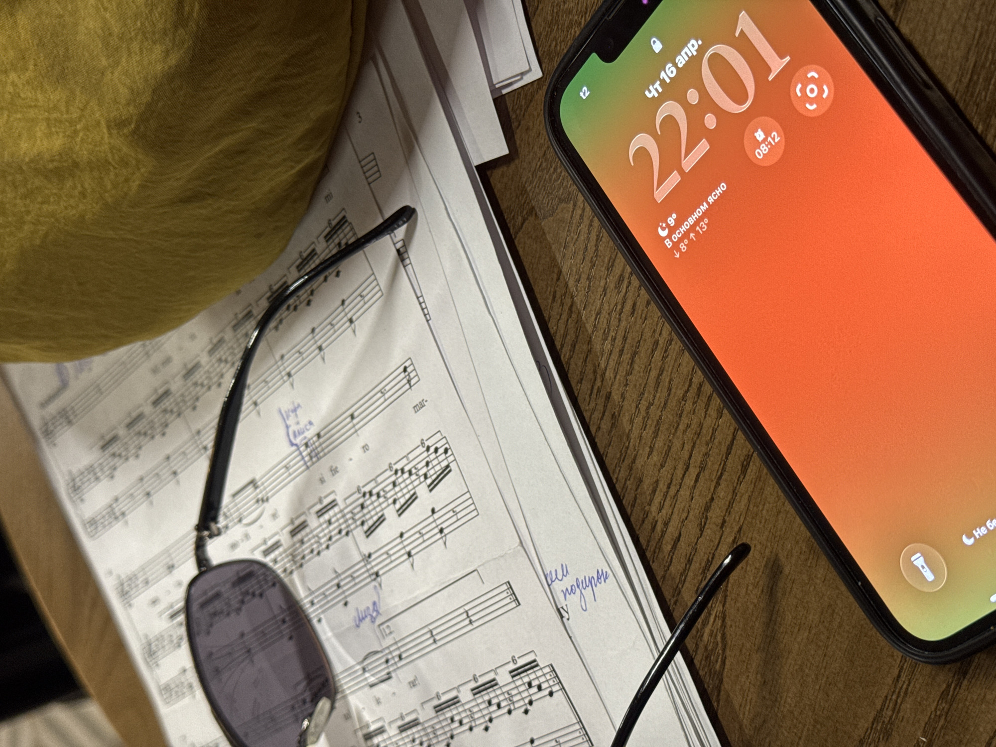 An overhead shot captures a collection of items resting on a wooden surface. The primary subjects include a smartphone, sheet music, sunglasses, and a textured yellow or mustard fabric object (possibly a cushion or garment).
The smartphone, positioned on the right, displays its lock screen with the time "22:01" (10:01 PM) and the date "Чт 16 апр." (Thursday, April 16). The screen also shows weather information in Russian: "9°" (9 degrees Celsius), "В основном ясно" (Mostly clear), and temperature range "↓ 8° ↑ 13°" (low 8°, high 13°). Another small time, "08:12", is visible, along with flashlight and camera icons, and the partial text "Не бе" (likely part of "Не беспокоить" - Do Not Disturb).
To the left, a pair of dark-lensed sunglasses with black frames lies on a stack of sheet music. The music sheets are filled with musical notation, including staffs, notes, and dynamics. Handwritten text in what appears to be Cyrillic script is present on the music, with visible words like "кем" and "подирок" (likely "подарок" - gift). The word "RAUCA" and fragments like "si fie" and "mar-" are also legible on the music. The yellow fabric object is partially visible in the top-left corner, and the wooden surface has a visible grain pattern. The scene suggests an indoor setting, possibly a desk or table, where someone might have been studying or playing music.