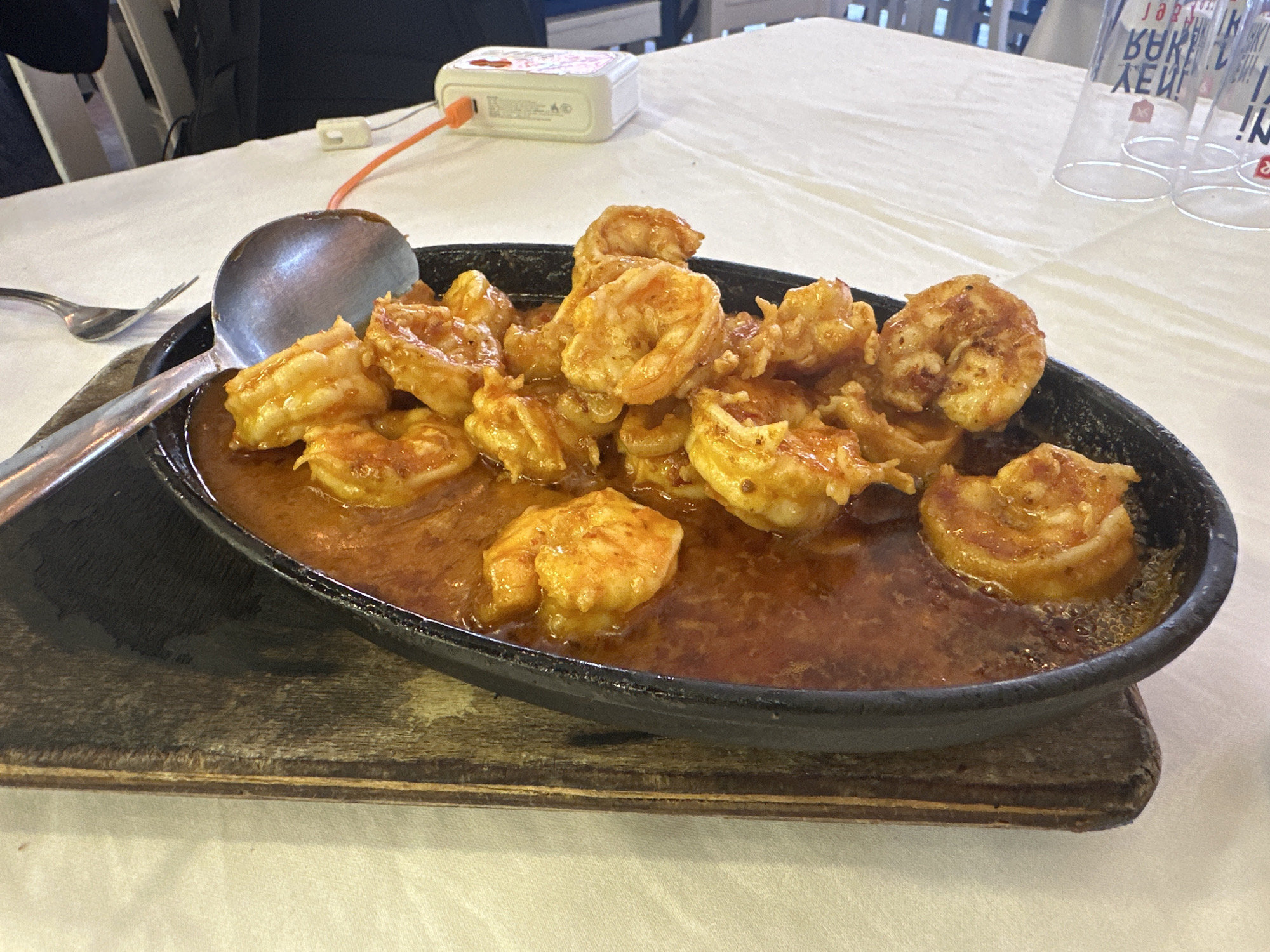 The image captures a culinary scene, specifically a dish of sautéed shrimp, served at a location within Fethiye, Turkey. The shrimp are arranged in a black, cast-iron skillet, submerged in a brownish-orange sauce, and set atop a dark, wooden board, presumably for presentation. The shrimp themselves are plump and coated in the same sauce. A large, stainless steel spoon rests to the side of the skillet, suggesting the dish is ready to be served.
The setting appears to be a restaurant or dining establishment, as indicated by the white tablecloth covering the table. In the background, there are glasses and other dining paraphernalia, along with a glimpse of a person's arm and shoulder, as well as a white charger-like object with an orange cord, presumably a phone charger. The lighting is bright and even, highlighting the textures and colors of the food and the surrounding objects. The overall impression is one of a casual, yet inviting, dining experience in Fethiye.