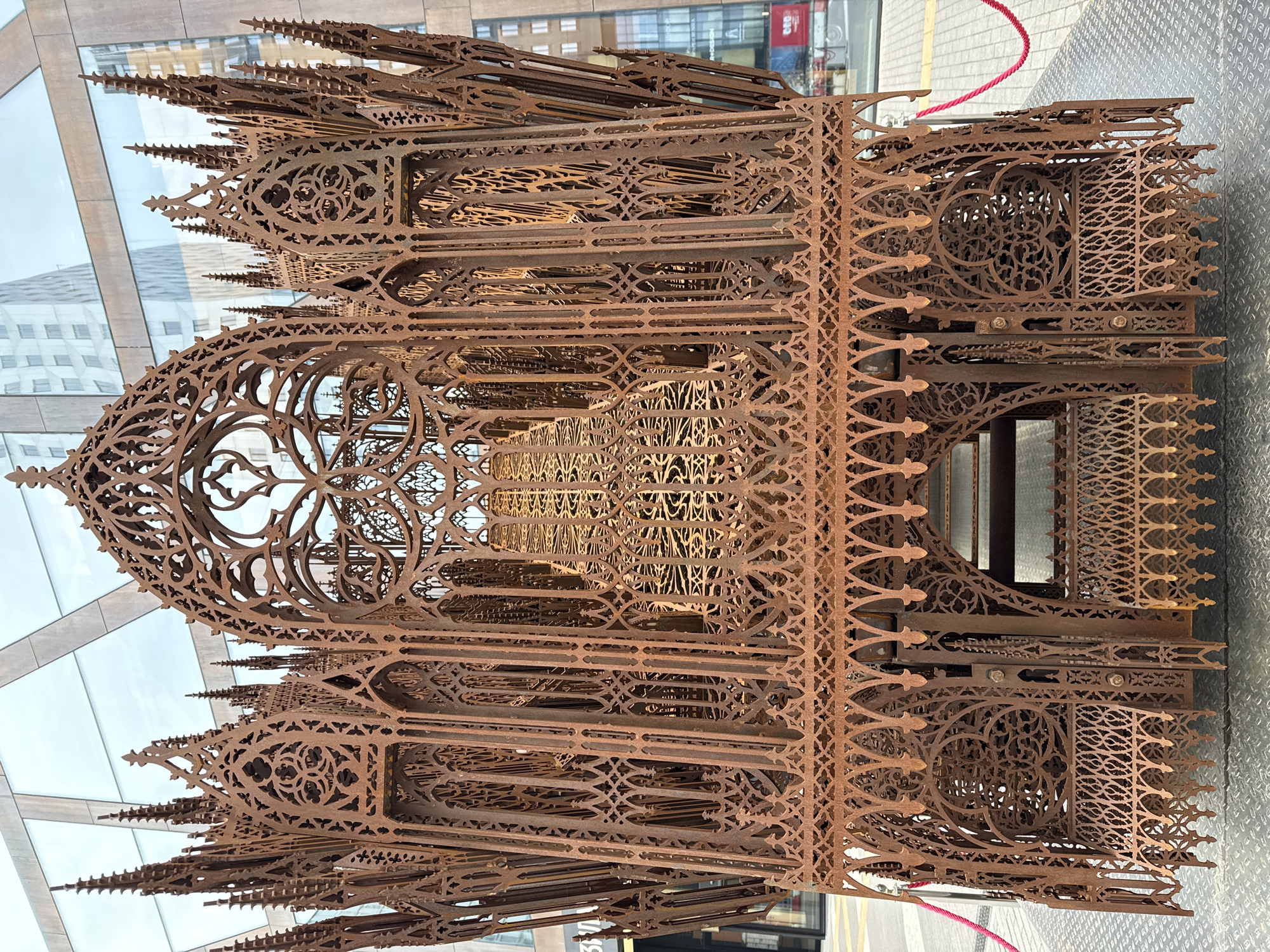 The image shows a detailed, rusted metal sculpture reminiscent of Gothic architecture, possibly a stylized cathedral facade. The sculpture is intricate, featuring pointed arches, tracery, and spires. It is located in an urban environment; modern glass buildings are visible in the background. A section of red rope is visible along the top right side of the sculpture. The sculpture is standing outside on a paved surface. The image suggests an outdoor art installation. The location is Moscow, Russia, based on visual cues in the background (such as the architectural style of the modern buildings and any identifiable Cyrillic text). No people are visible in the image.