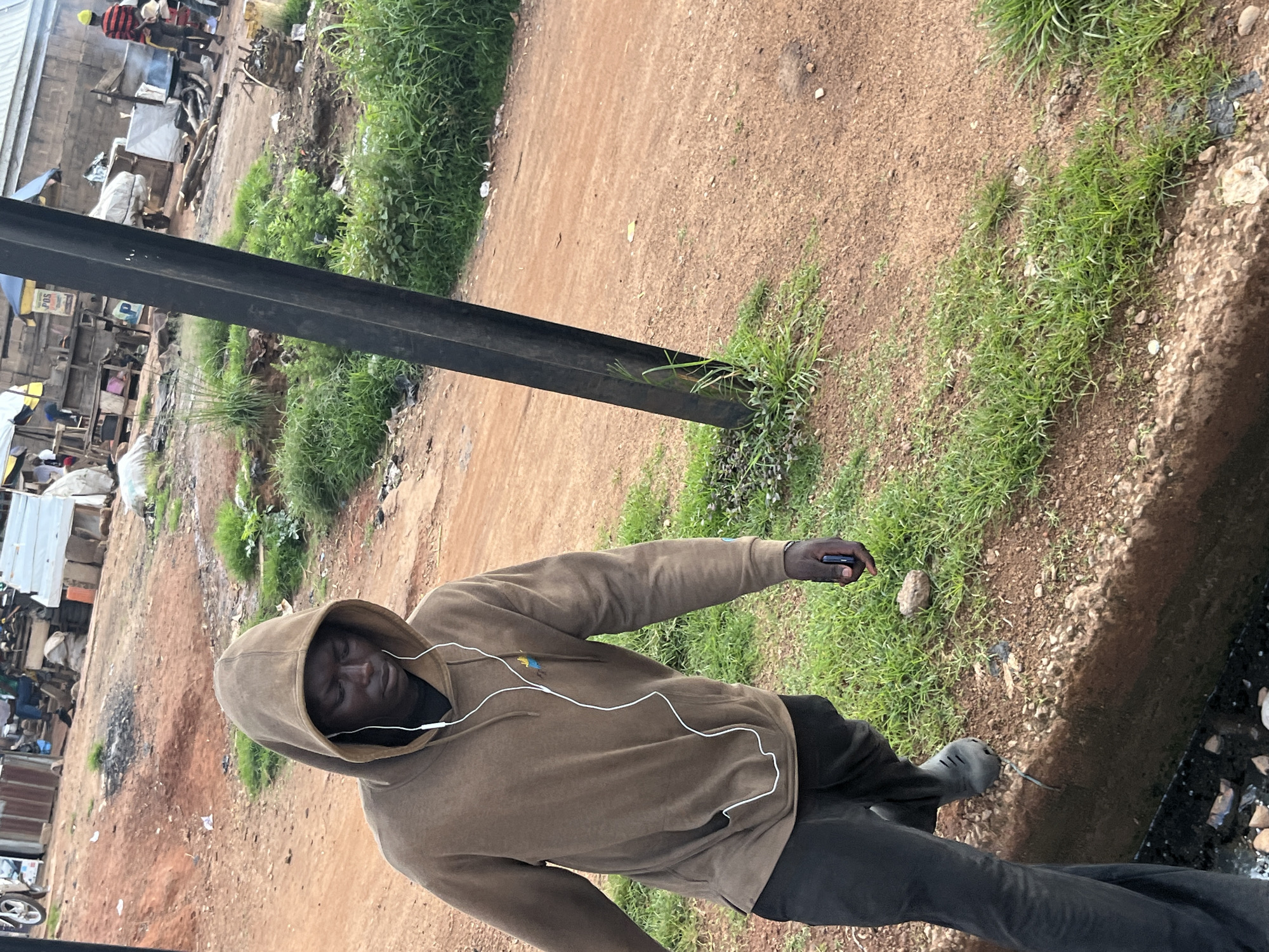 The image shows a young Black male wearing a brown hooded sweatshirt and dark pants, standing outdoors with earbuds in. He is holding a dark-colored smartphone in his right hand. The background suggests a somewhat unkempt, rural or informal urban setting with dirt ground, scattered debris, and patches of green grass and weeds. In the distance, there are some makeshift structures that could be stalls or workshops, and hints of other people in casual attire, possibly engaged in commerce or daily activities. The overall impression is of a casual, everyday moment in a developing area. The light suggests it is daytime. There are no clear indications of the weather, but the presence of grass implies it's not severely dry. The scene appears to be in Gura Top, Nigeria.