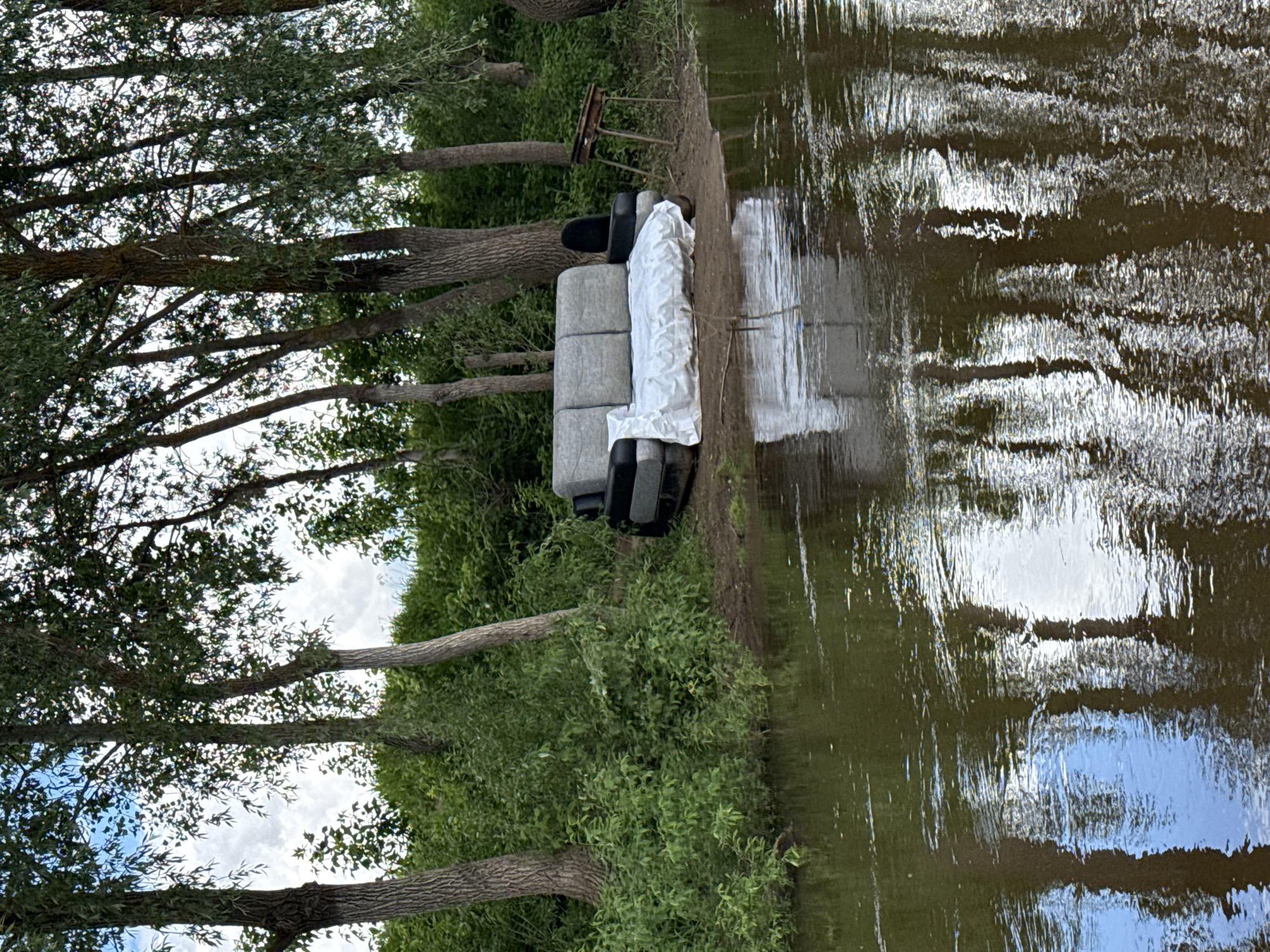 The image shows a discarded grey sofa partly covered with a white sheet, positioned on the riverbank in Izh-Boyki, Russia. Next to it is an old chair. The surrounding environment includes trees, bushes, and the water of the river. The reflection of the sky is visible in the water.