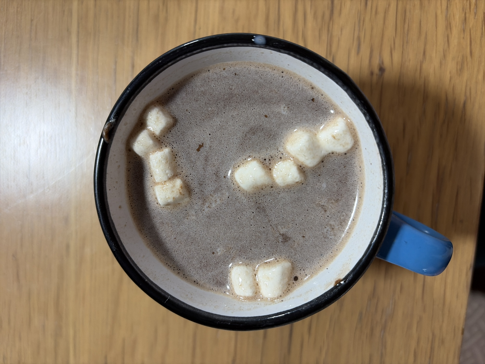 This is a top-down, eye-level shot of a mug of hot chocolate with marshmallows on a wooden surface. The mug is white with a black rim and a bright blue handle. The hot chocolate is foamy and topped with several white marshmallows, some of which are partially submerged. The wooden surface appears to be a table with visible grain. The image captures a close-up view of the mug, with the background of the wooden table slightly out of focus. No people are visible in the image. The location of Oxentea, Moldova, is not directly evident from the visual information provided in the image.