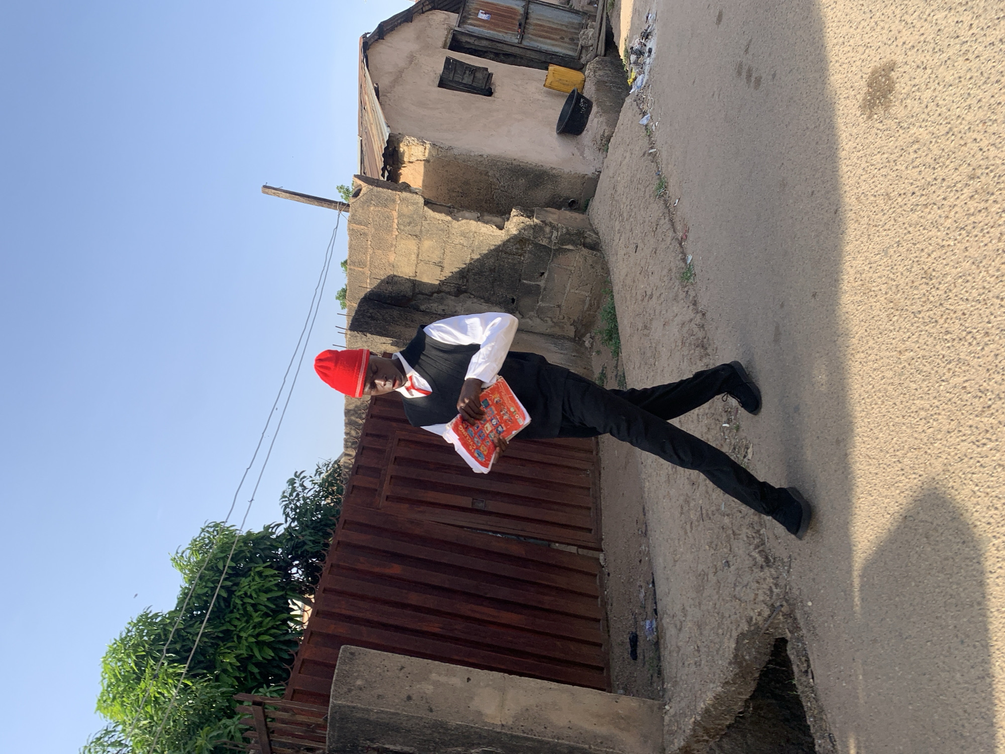 A man wearing a red cap, white shirt, black vest, and black pants stands on a street in Jos, Nigeria. He is holding a colorful object with his hands. The street is paved. Behind the man is a building with a weathered exterior and a corrugated metal roof. Adjacent to the man is a metal gate or fence. Trees and vegetation are visible on the left side of the image. The sky is clear and blue. The photo appears to be taken during the daytime.