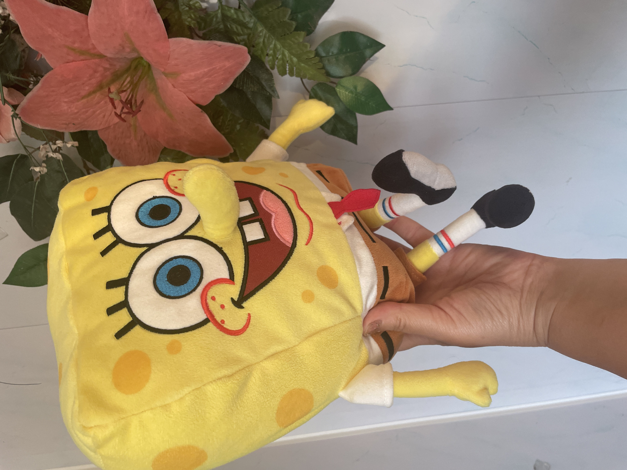 A close-up, eye-level shot shows a stuffed Spongebob Squarepants character held by a person's hand against a light blue tiled background. The Spongebob plush is predominantly yellow with characteristic brown square pants, a white shirt, and a red tie. Its large blue eyes and wide smile are detailed with black stitching. To the right of the Spongebob plush, a large, bright pink lily flower and green foliage are visible. The person's hand, which has neatly manicured nails, is positioned to support the Spongebob plush's midsection and lower body. The background appears to be a wall with a subtle, marbled pattern. The lighting suggests an indoor setting, possibly a home. There is no visible indication of the city of Shpykolosy, Ukraine.