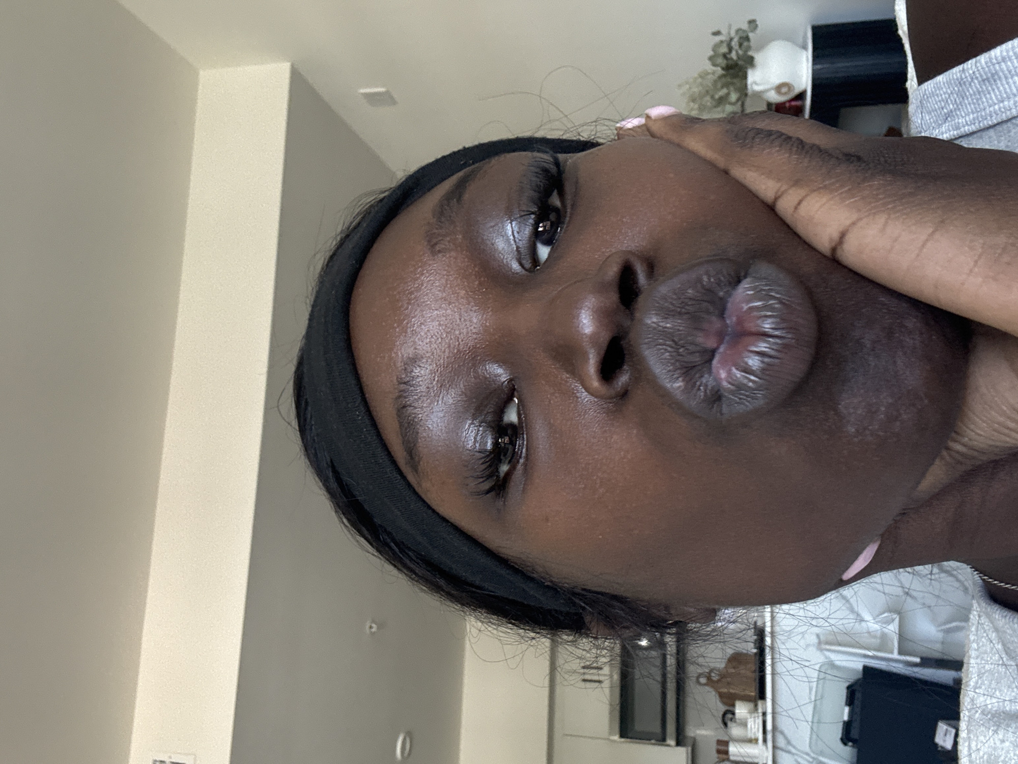 A close-up, rotated image shows a Black woman with dark skin, wearing a black headband. Her left hand touches her face. She is making a kissing face. The background suggests an interior space, possibly a modern apartment or home. Specific details such as furniture and lighting fixtures indicate a domestic setting within a city apartment. It's not possible to confirm the location is in Los Angeles without further contextual details.