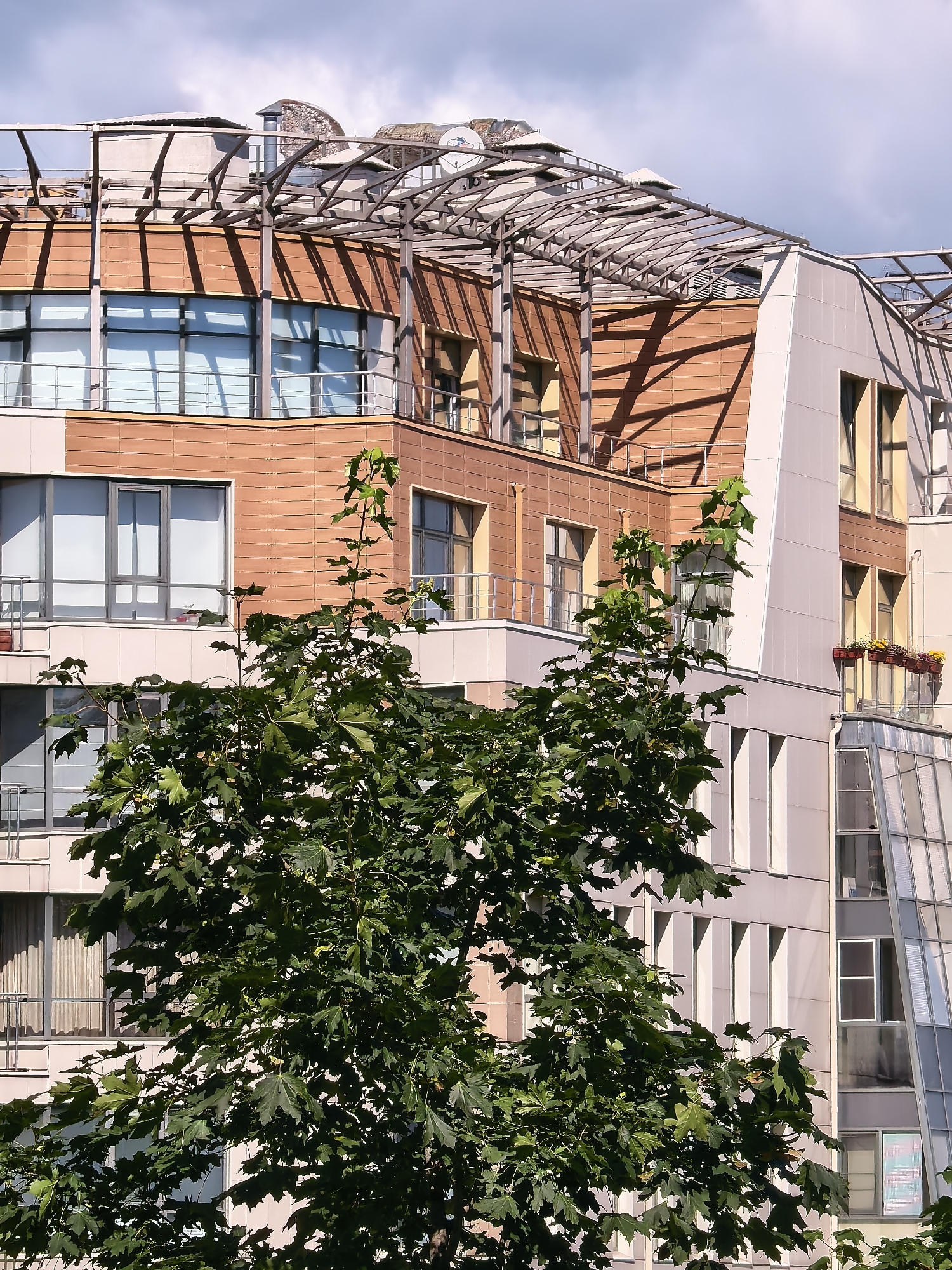 This is a medium-shot, eye-level, outdoor photograph of a modern building in Sestroretsk, Russia, on a sunny day with a partially cloudy sky. The building features a multi-faceted design with a mix of tan, cream, and glass facades. A prominent architectural element is a metal canopy structure extending over the upper floors, casting shadows. Several windows, some with balconies, are visible. In the foreground, a large tree with lush green leaves partially obscures the lower and middle sections of the building. The overall impression is of an urban residential or commercial structure integrated with natural elements.