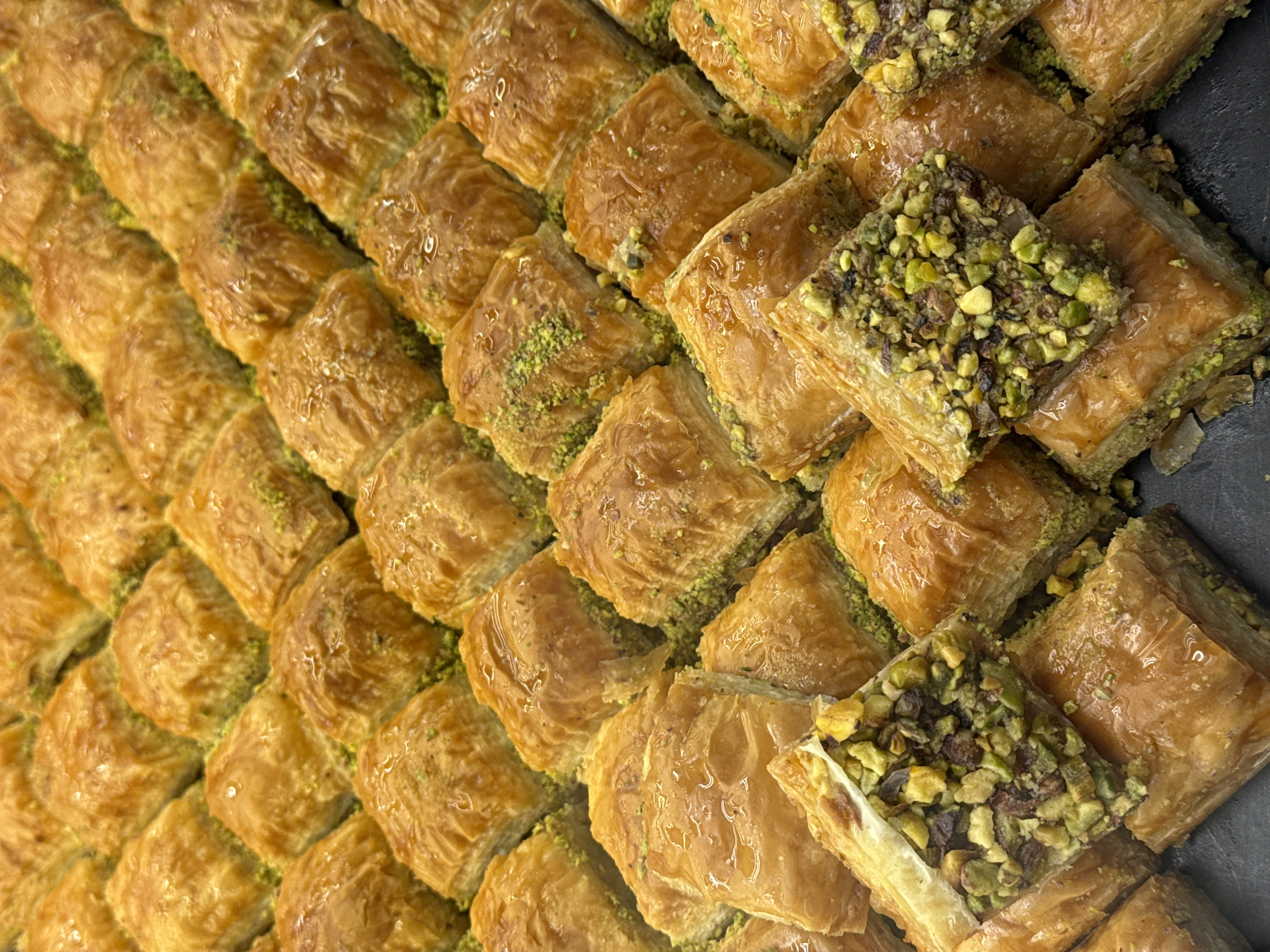 The image shows a close-up, overhead view of a large tray of freshly baked baklava, a traditional Middle Eastern and Balkan pastry. The baklava pieces are diamond-shaped and arranged in neat rows. They have a golden-brown, flaky crust, glistening with a syrup. Many of the pieces are topped with finely chopped pistachios, visible as a vibrant green dusting. One piece in the foreground is broken open, revealing layers of thin pastry and a filling of nuts and possibly other ingredients. The baklava is displayed on a dark, flat surface, likely a tray or countertop. The lighting is bright, highlighting the textures and shine of the pastries. The setting is not specified as being in Arnavutköy, Turkey, nor is there any information about people, actions, or interactions.