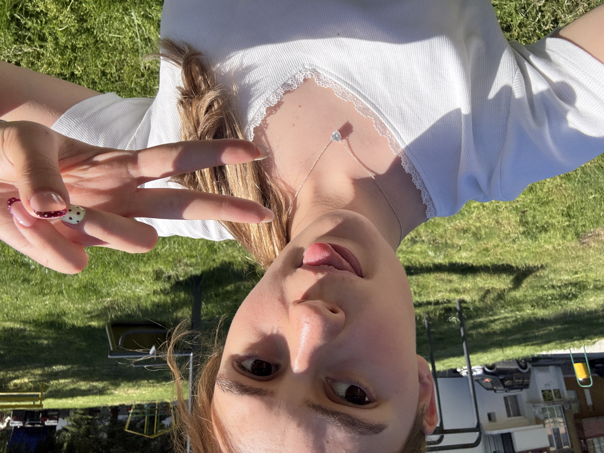 A young woman is lying on green grass in Panagyurski kolonii, Bulgaria. She is wearing a white top with lace trim around the neckline and a silver necklace with a pendant. She is holding up her left hand with her fingers in a peace sign, showing two manicured nails with decorative patterns. Her tongue is sticking out slightly. In the background, there are trees, playground equipment, a house, and cars.