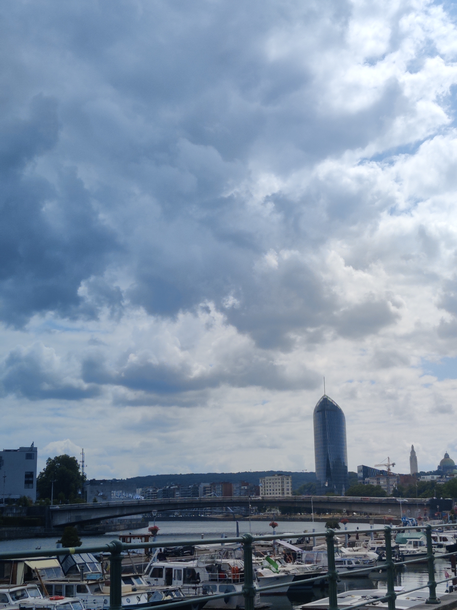The image captures a view of the Meuse River in Liège, Belgium, under a cloudy sky. In the foreground, several boats are moored along the riverbank. A metal railing runs across the bottom of the frame, partially obscuring the boats. The middle ground features a bridge spanning the river, with traffic visible on it. Beyond the bridge, a distinctive curved skyscraper, the KBC Tower, stands tall in the distance. Several other buildings and trees are scattered across the cityscape. In the background, rolling hills can be seen. The overall atmosphere is overcast, suggesting a potentially inclement weather day.