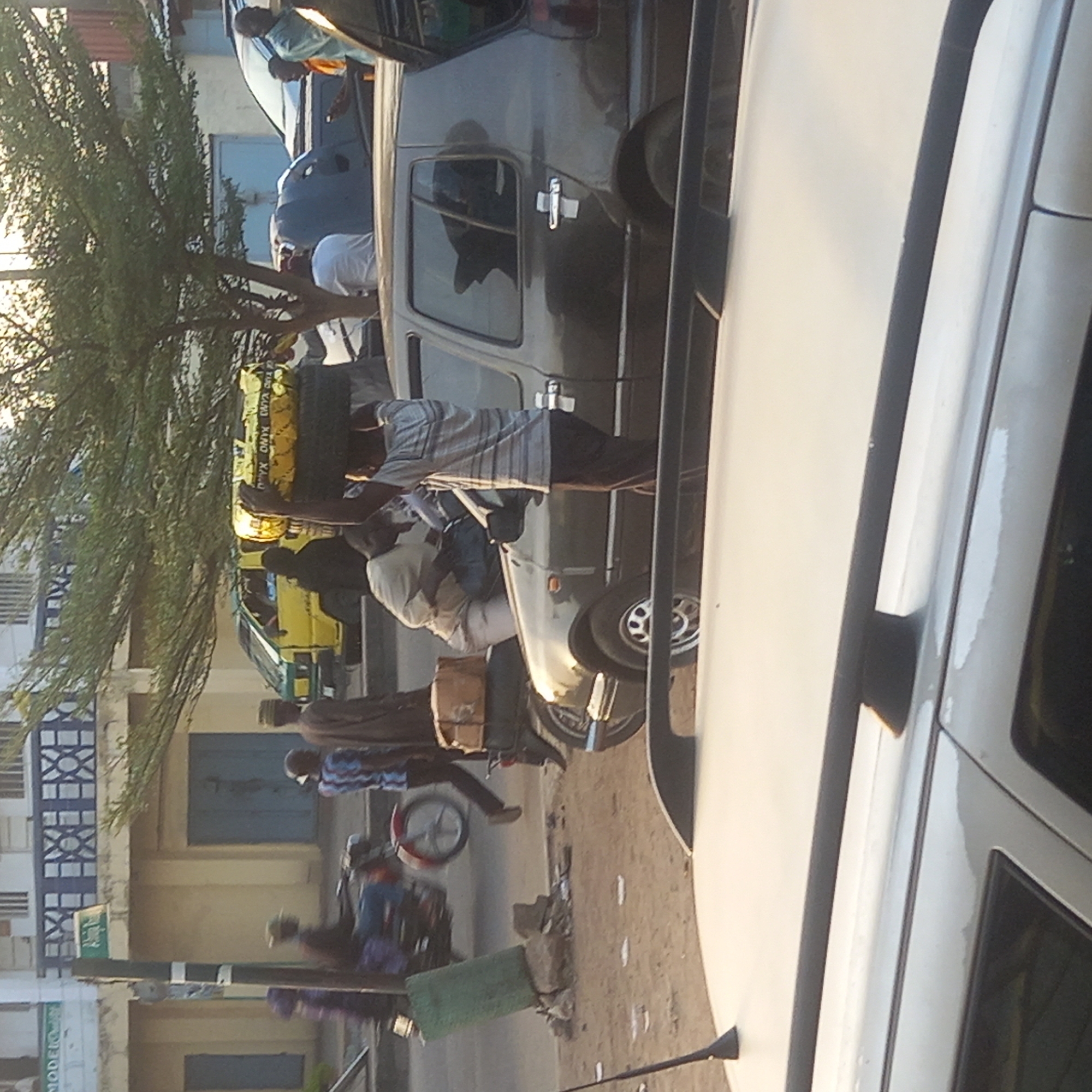 The image captures a bustling street scene in Sardauna, Nigeria. Several individuals are visible, including a man carrying goods on his head, and another man pushing a cart. Two men on a motorcycle. There are several cars parked along the roadside. The environment includes buildings, trees, and what appears to be a dirt or gravel road.