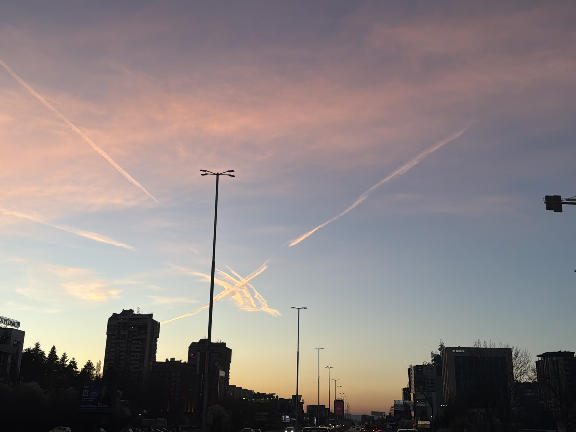 The image depicts an urban setting at dusk, likely in Sofia, Bulgaria, given the architectural styles and street lighting. The sky transitions from pink to blue, marked by several contrails from aircraft. Foregrounded are silhouettes of buildings, trees, and streetlights along a busy thoroughfare with visible traffic. A traffic light is partially visible in the right of the frame.