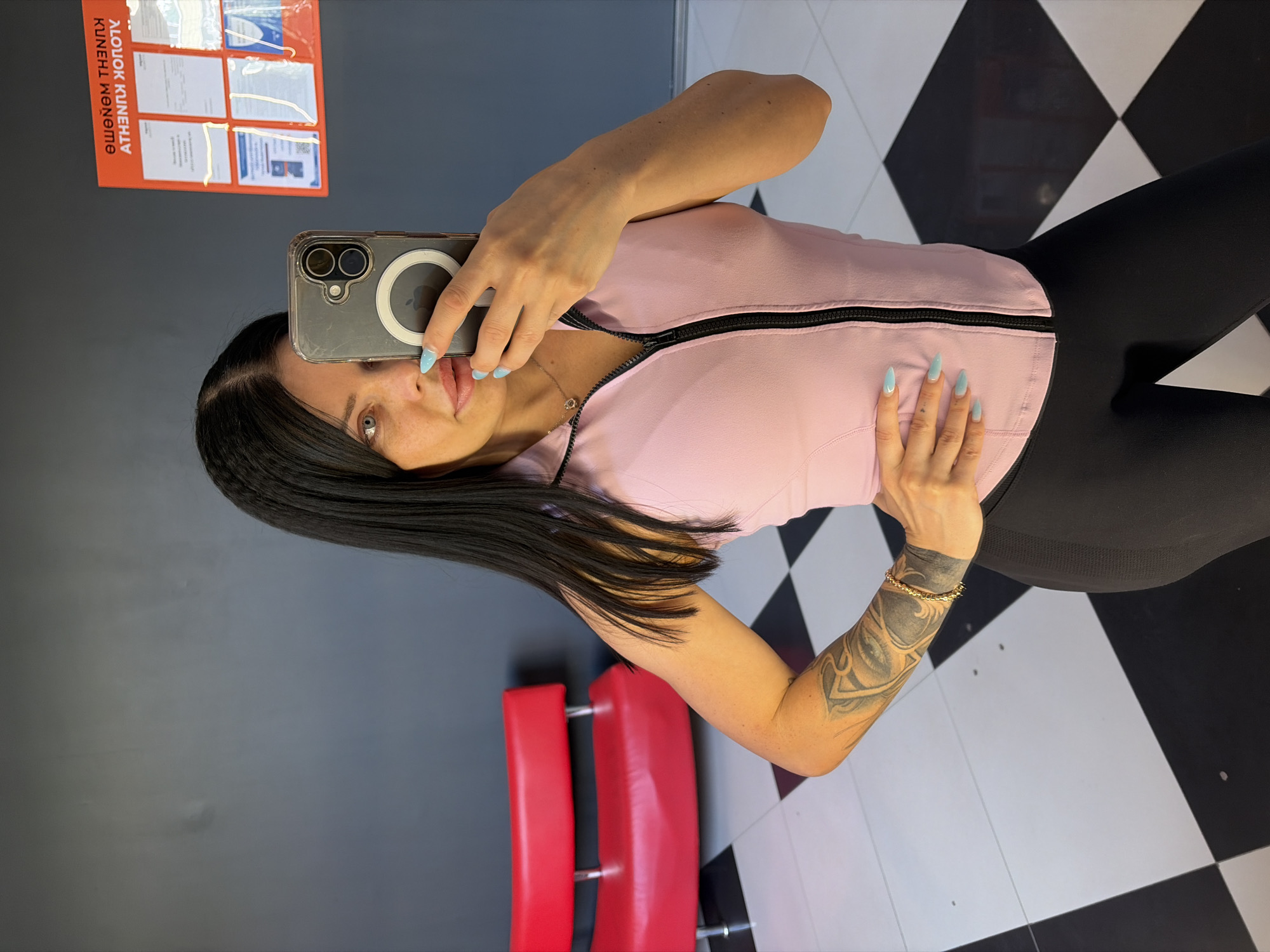 A woman is taking a mirror selfie in what appears to be a gym or workout space. She is wearing a light pink zip-up top and black leggings. Her hair is long and dark, and she has light blue nail polish on. She is holding a smartphone in her right hand, which is angled to capture her reflection. Her left arm is bent, and her hand rests on her chest, displaying a tattoo on her forearm and a bracelet on her wrist.
In the background, a red piece of gym equipment, possibly a bench or chair, is visible. The floor has a black and white checkered pattern. To the left of the woman, there is a gray wall with an orange poster attached to it. The poster displays several documents or certificates in clear plastic sleeves. Some text is visible on the poster, but it is not fully legible. The lighting in the image suggests an indoor setting, likely during the day. The woman's expression is neutral, and the overall tone of the image is casual and personal.