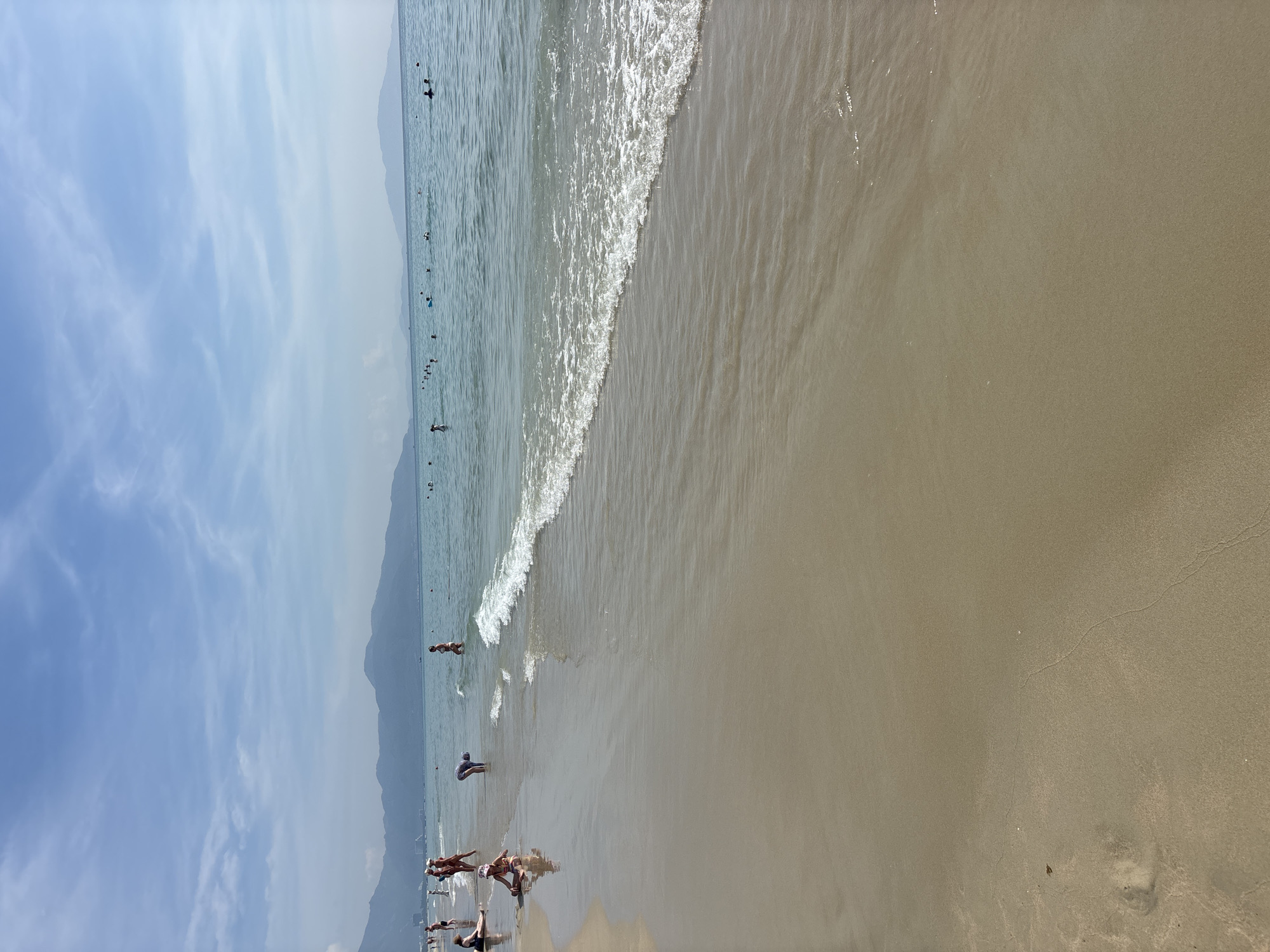 The image depicts a broad, sandy beach with a gentle shoreline leading into the ocean. The water is calm, with small waves lapping the shore, and the sky above is a clear blue with scattered white clouds. Several people are visible on the beach and in the water, some standing, some sitting, and others wading or swimming. The distant horizon is marked by a chain of mountains, suggesting a coastal landscape. The overall scene conveys a relaxed atmosphere of a summer day at the beach.