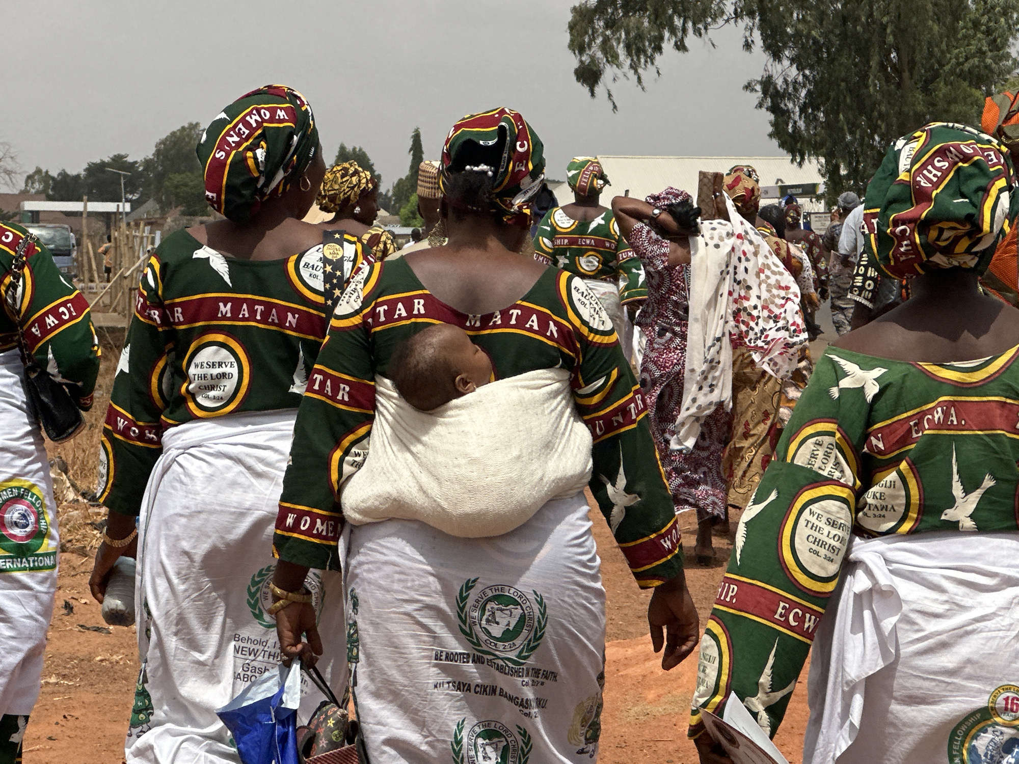 A medium shot shows a group of women walking in an outdoor area, possibly in the city of Unknown, Unknown. The women are wearing matching outfits that include green, maroon, and gold patterned tops with a white skirt, and similarly patterned head coverings. The tops have inscriptions and symbols, including doves and phrases such as "We Serve The Lord Christ." One of the women in the foreground carries a baby in a sling on her back. Some of the women carry items such as an umbrella, a water bottle, and papers. Other people are visible in the background, and there are trees and buildings in the distance. The ground appears to be reddish dirt.