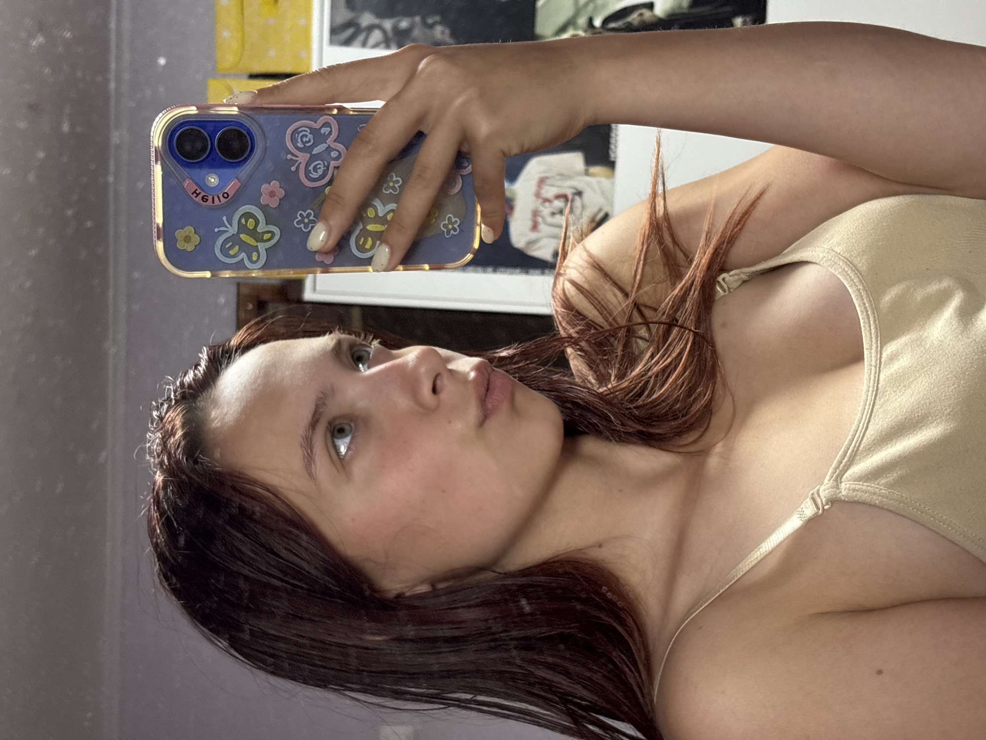 A young woman with long, dark red hair is taking a mirror selfie. She is wearing a beige tank top. Her right hand is holding a smartphone with a purple case adorned with butterflies and flowers. The phone is partially obscuring her face. She is looking at the phone's screen. The background appears to be a room with a mirror and posters on the wall. The lighting suggests it may be indoors, possibly at night or in a dimly lit space. There is no visible indication of the location being in Прикамский, Russia, nor any details that would place it in a city.