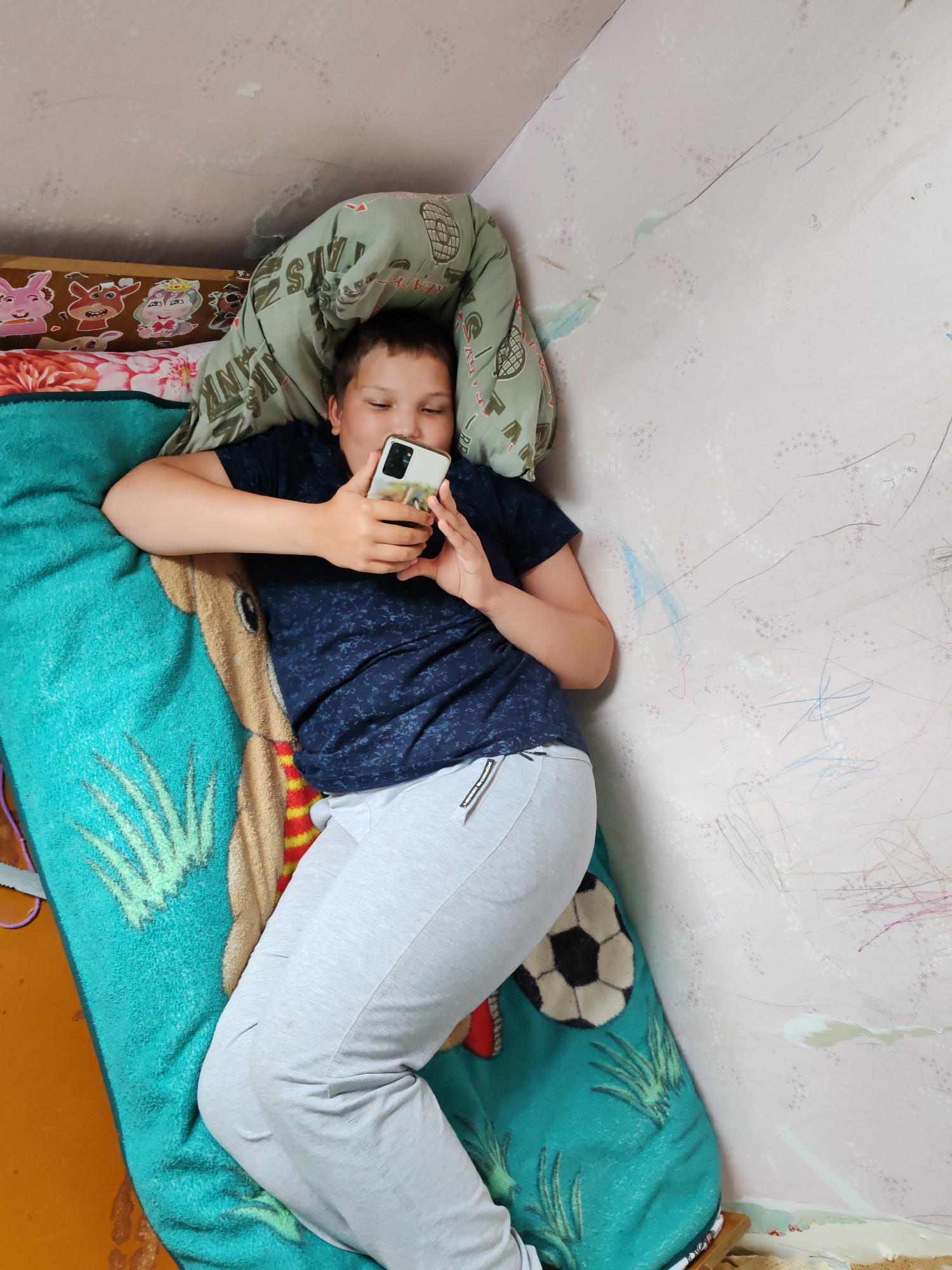 A boy lies on a bed in a room. He is holding a smartphone and looking at the screen. The room has light pink walls with crayon marks. The bed is covered with a green and brown blanket. A soccer ball is on the bed. This is taken in the city of Чухлэм, Russia.