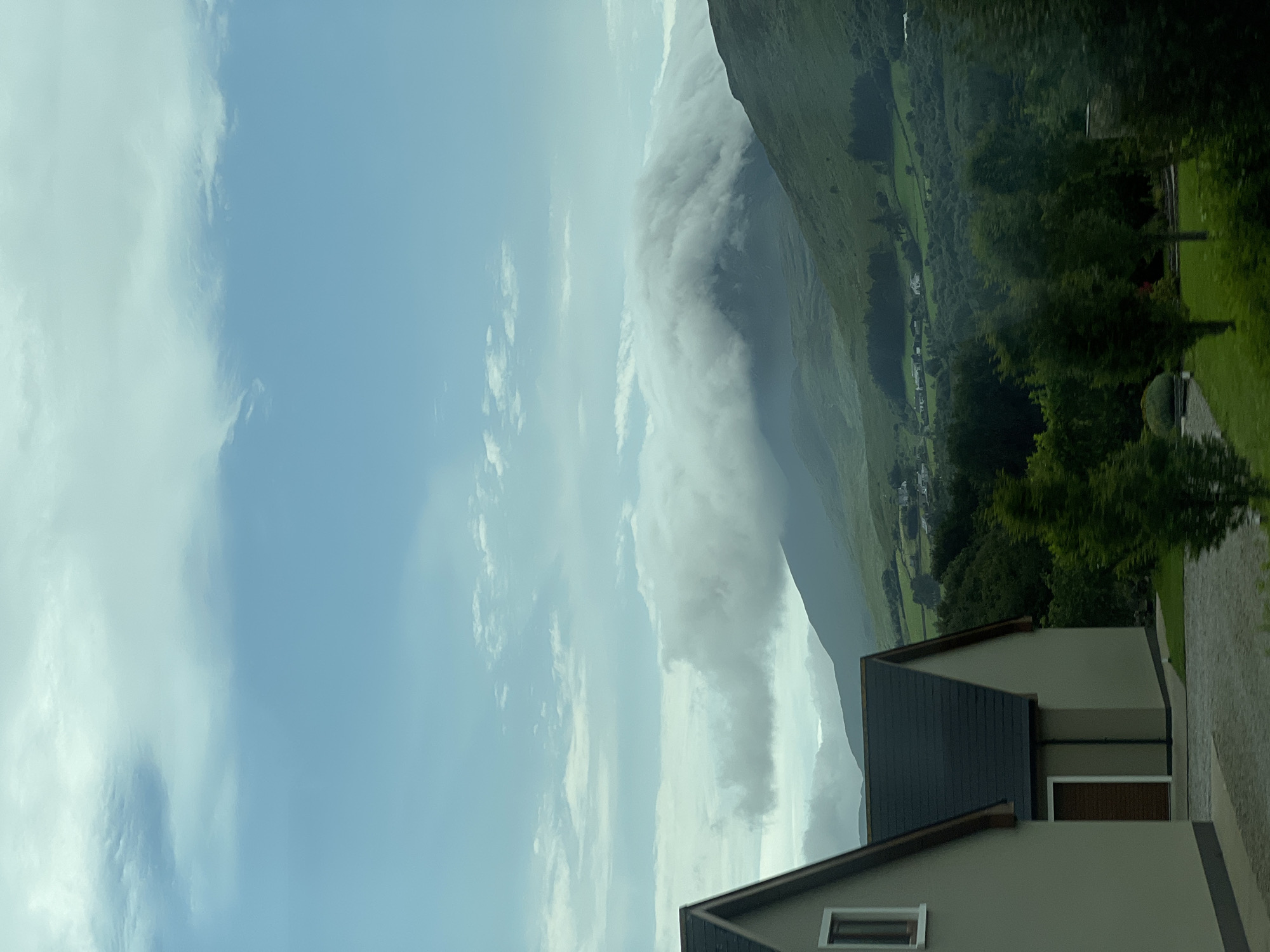 The image depicts a cloudy sky over a lush green mountainous landscape. In the foreground, two modern houses with grey facades and dark roofs are visible on the left side. The houses appear to be residential properties. The background features rolling hills covered in vibrant green vegetation, dotted with scattered white houses. A large mountain in the mid-ground is partially obscured by dramatic, low-hanging clouds. The overall scene suggests a rural or semi-rural setting. The location is identified as Creeslough, Ireland. There are no people visible in the image.