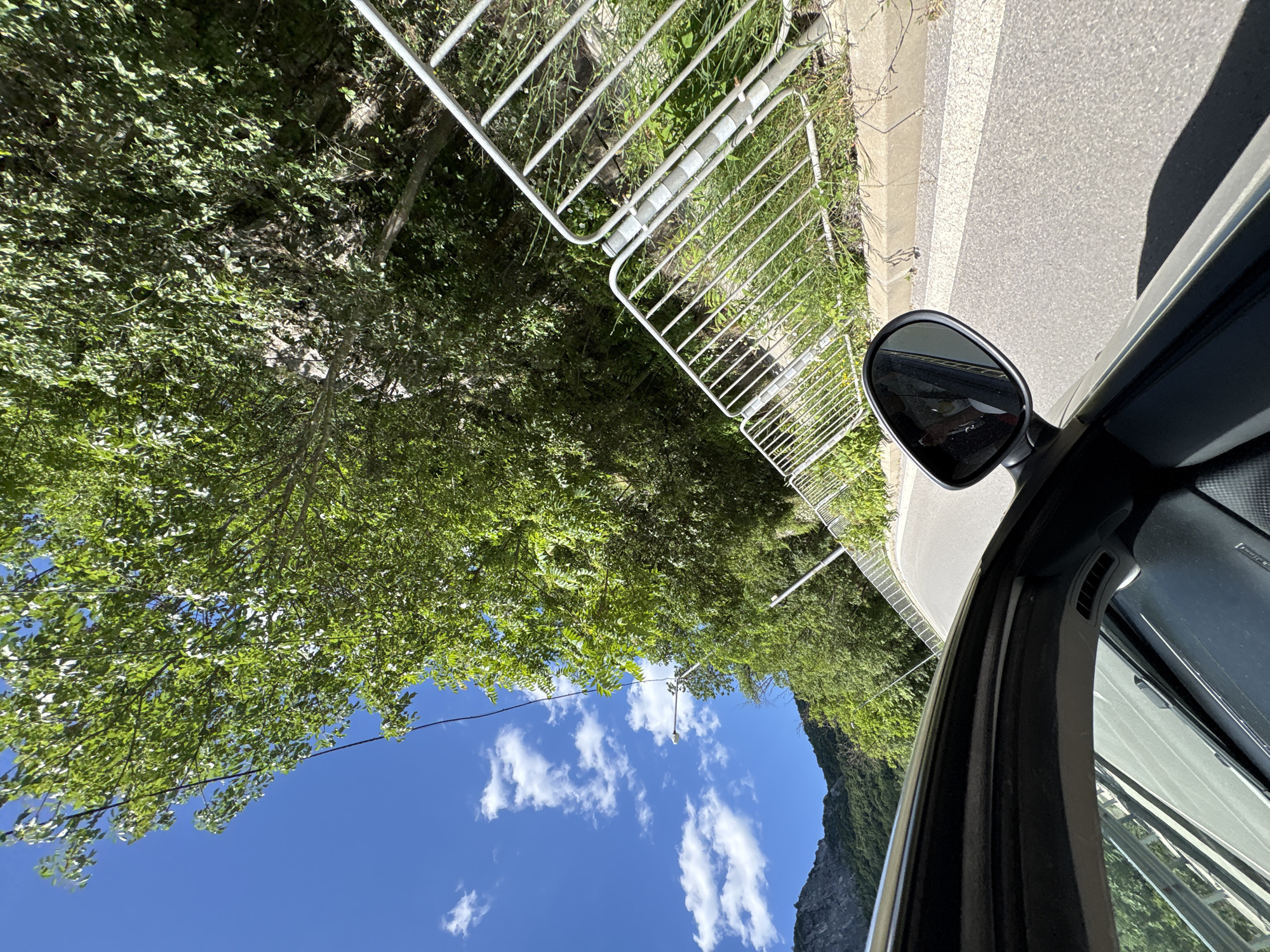 The image depicts a view from the interior of a car looking out a side window and rearview mirror while driving on a paved road. The road curves to the right and is bordered by a metallic guardrail on the right side. Beyond the guardrail, there is dense green foliage and trees, indicating a natural, possibly mountainous or hilly environment. In the distance, on the left, a steep, rocky mountain slope covered with trees is visible under a bright blue sky with scattered white clouds. A power line is visible crossing the sky and draped over tree branches. The car's interior is partially visible in the foreground, with a portion of the door panel and dashboard. The rearview mirror reflects a person or persons within the car. The scene appears to be captured during daylight, likely late morning or early afternoon, given the brightness and shadows. The specific location in Vratsa, Bulgaria, is not identifiable from the visual elements alone, but the landscape suggests an area outside the city center, possibly along a scenic route or on the outskirts leading towards mountainous terrain.