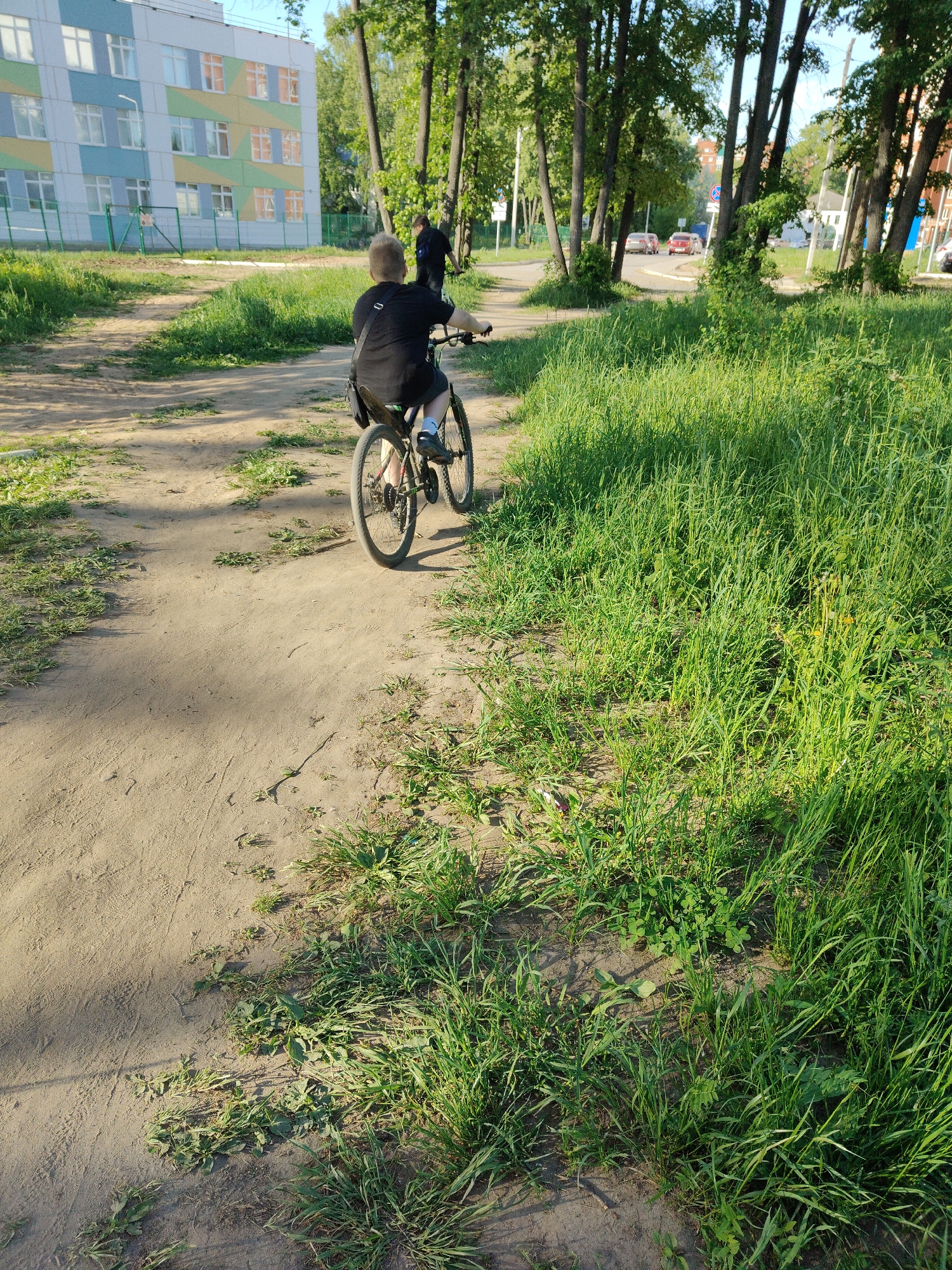 A boy is riding a bicycle on a dirt path, heading away from the camera, in Unknown, Unknown. The path is bordered by tall green grass on the right and a building in the background on the left. There are trees alongside the path and another person is walking in the same direction as the boy on the bike. In the far distance, cars can be seen on a road.