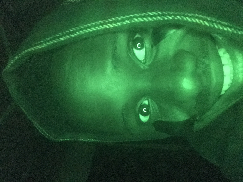 A person's face is illuminated in green, possibly through night vision. The face is upside down and wears a hooded garment with a stitched edge. The eyes reflect light, each appearing to have a letter "C" superimposed on the pupil. The person is smiling, revealing their teeth. The background is dark and obscured.