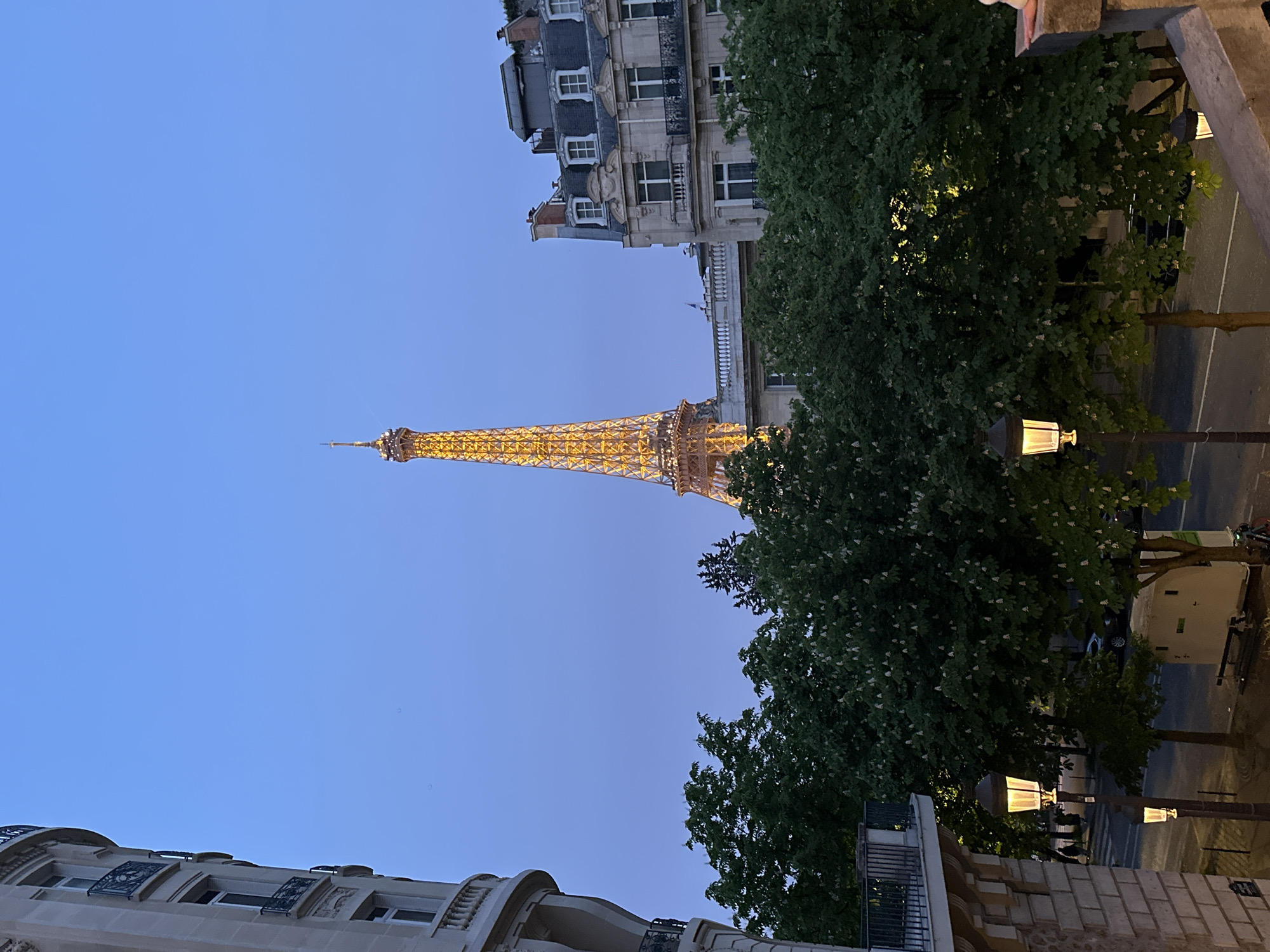 The image shows the Eiffel Tower in Paris, France, lit up at dusk. It is framed by buildings and trees. A street with lampposts is visible on the right side of the image.