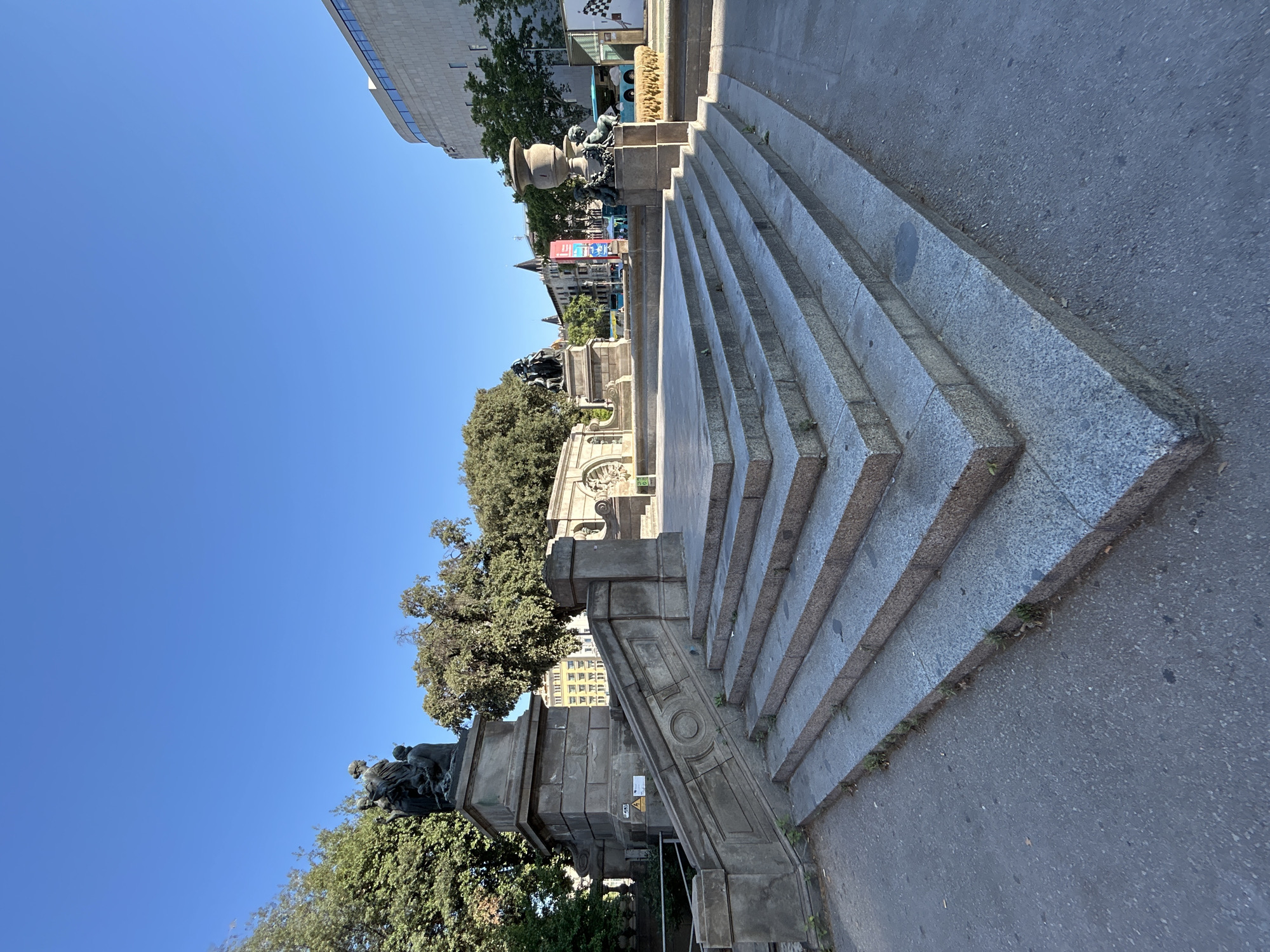 The image captures a public monument on a bright, clear day. The monument appears to be made of stone and features intricate carvings and statues. A wide set of granite stairs leads up to the monument, with a paved walkway on the right.
In the background, a modern building with a glass facade is visible, contrasting with the older architectural style of the monument and surrounding older buildings. Trees with lush green foliage frame the monument, suggesting a park or public square setting. The sky is a deep, cloudless blue, indicating fair weather and likely midday.
While no specific event is depicted, the monument suggests it is a place of historical or cultural significance. The overall atmosphere is serene and still, with no people actively engaged in the scene. The location is identified as Barcelona, Spain, which, combined with the architectural style, hints at a European urban environment. Some caution signs are visible on the side of the monument, one indicating a danger of electricity. The clear, bright lighting suggests it is daytime, likely morning or afternoon.