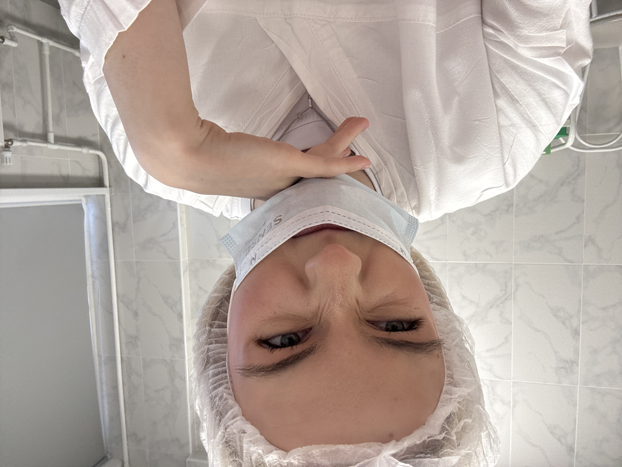 A selfie shot of a person wearing a white surgical cap and a blue surgical mask. The person is also wearing a white garment, possibly a medical coat. The background appears to be a tiled wall, possibly in a clinical or hospital setting. The person is positioned at an angle, looking down towards the camera. The exact location in Saint Petersburg, Russia, cannot be determined from the image.