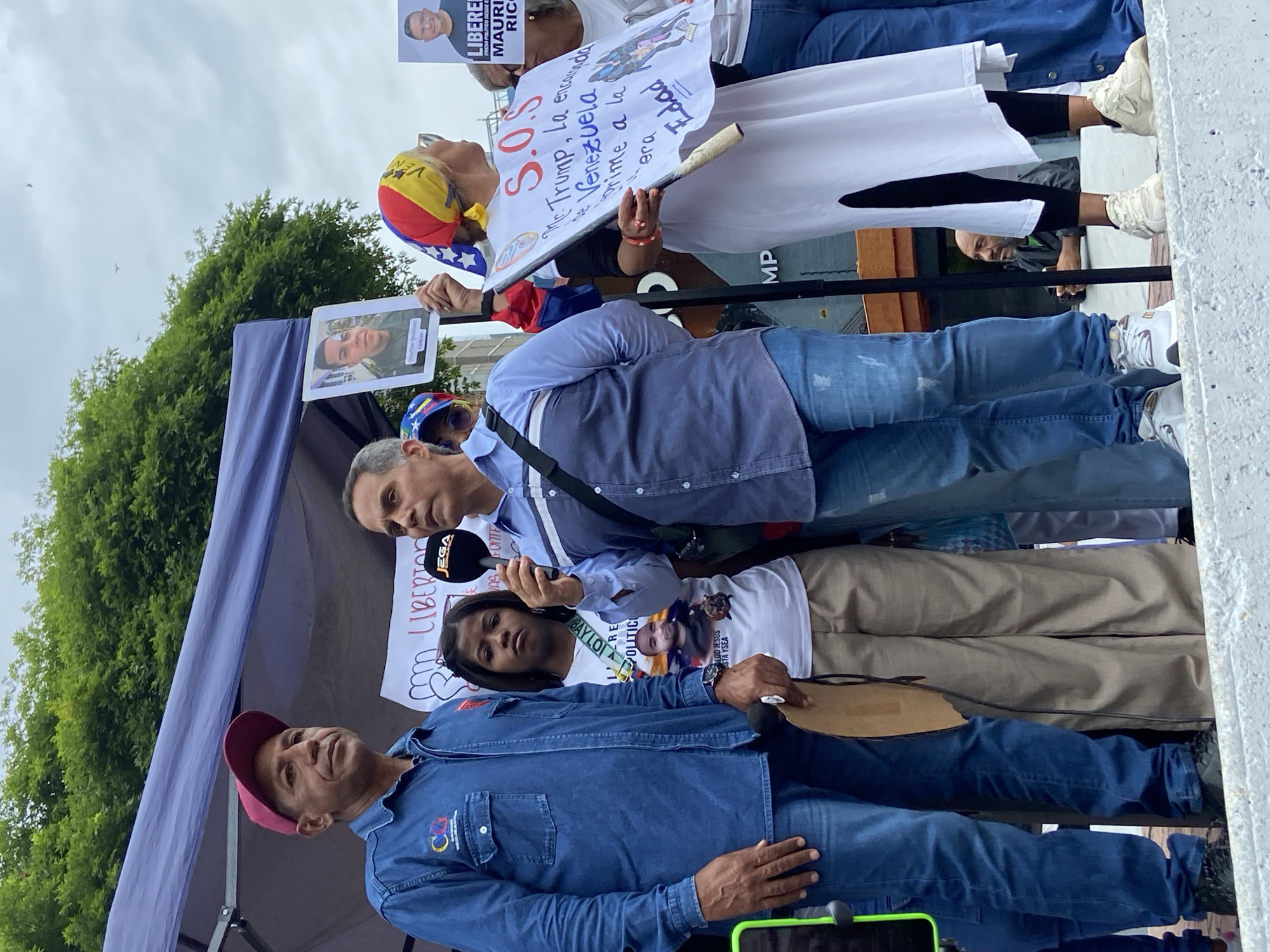 This image captures an outdoor political protest or rally in Caracas, Venezuela, under an overcast sky. Several individuals are present, advocating for various causes. In the foreground, a man in a blue denim shirt and a red cap stands, looking to his right. To his left, another man, wearing a light blue shirt and jeans, is speaking into a microphone with a "JEGA" logo, indicating he might be addressing the crowd or media. Behind him, a woman with dark hair wears a white t-shirt featuring text including "LIBERTAD" and "JESUS YSEA," along with a photo of a man.
To the right of the speaker, a person with a bandana in the colors of the Venezuelan flag (yellow, blue, red) and sunglasses holds a white sign. The sign, partially obscured, reads "S.O.S Mr. Trump, La encarcelada de Venezuela primer a la era edad," indicating an appeal to former US President Donald Trump regarding the situation in Venezuela. Other signs and posters are visible, including one with a man's face and the text "LIBEREN PRESO POLÍTICO DESDE EL MAURICIO RICARDO" (Free Political Prisoner Mauricio Ricardo), and another with a man's face identified as "Jonathan Quintero Quevedo." A blue canopy structure is visible on the left, with green trees in the background, suggesting a public park or street setting. The overall mood appears serious and determined, reflecting the nature of a political demonstration.