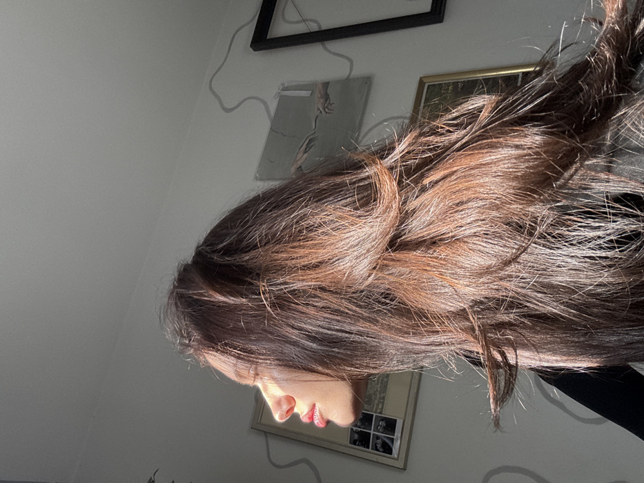 The image displays a female individual, partially visible in profile, indoors in Tel-Aviv, Israel. Her face, oriented towards the left, is illuminated by strong natural sunlight, highlighting her left cheek, nose, and lips, which exhibit a reddish hue. Her eyes appear closed or directed into the light. Her dark brown hair, featuring reddish highlights, is layered and cascades down her back, catching the sunlight. She is dressed in dark clothing, possibly a long-sleeved top. The background consists of a light-colored wall. On the left, a light-framed picture containing multiple small black-and-white photographs is mounted. Towards the center-right, a print depicting Michelangelo's "The Creation of Adam" is affixed to the wall. A section of a dark-framed object is visible on the far right. A grey electrical cable runs in a wavy pattern across the wall, passing behind the "Creation of Adam" print. The photograph is taken from a low, upward-looking angle.