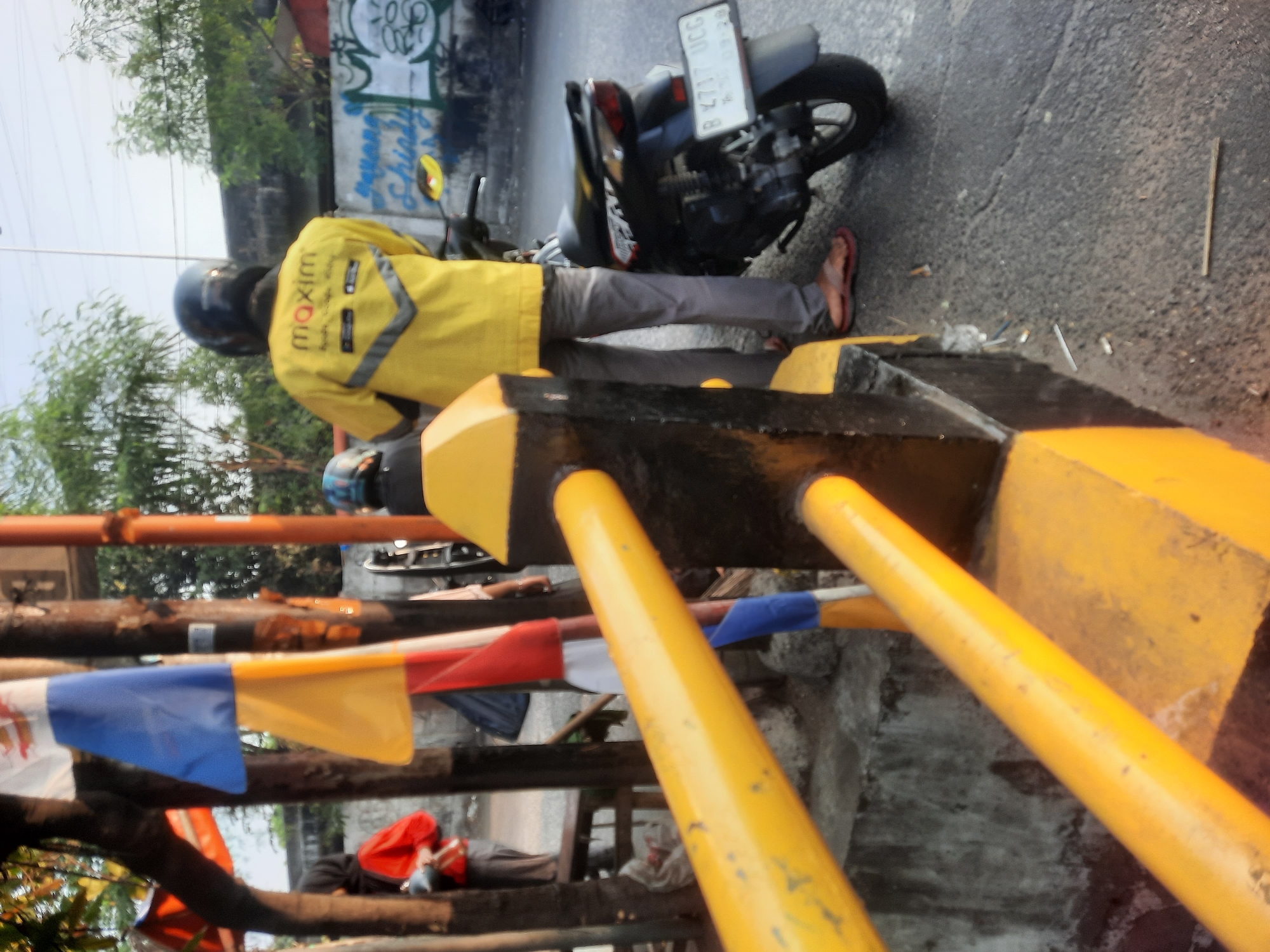 The image depicts an outdoor scene in North Jakarta, Indonesia, possibly along a street or roadside. In the foreground, a bright yellow barrier with black accents is visible, with a blurred perspective suggesting it's a close-up shot of the barrier itself. Behind the barrier, a person wearing a yellow jacket with "maxim" branding and a black helmet is standing next to a black motorcycle. The person is wearing grey pants and brown sandals. The motorcycle has a license plate that reads "B 7117 UCG". In the background, there are trees, buildings, and graffiti on a wall. Several flags, including blue, yellow, and red ones, are hanging from poles. Another person in a red jacket and black hijab is partially visible on the left side of the image, standing near a wooden bench. The overall impression is of a busy urban environment.