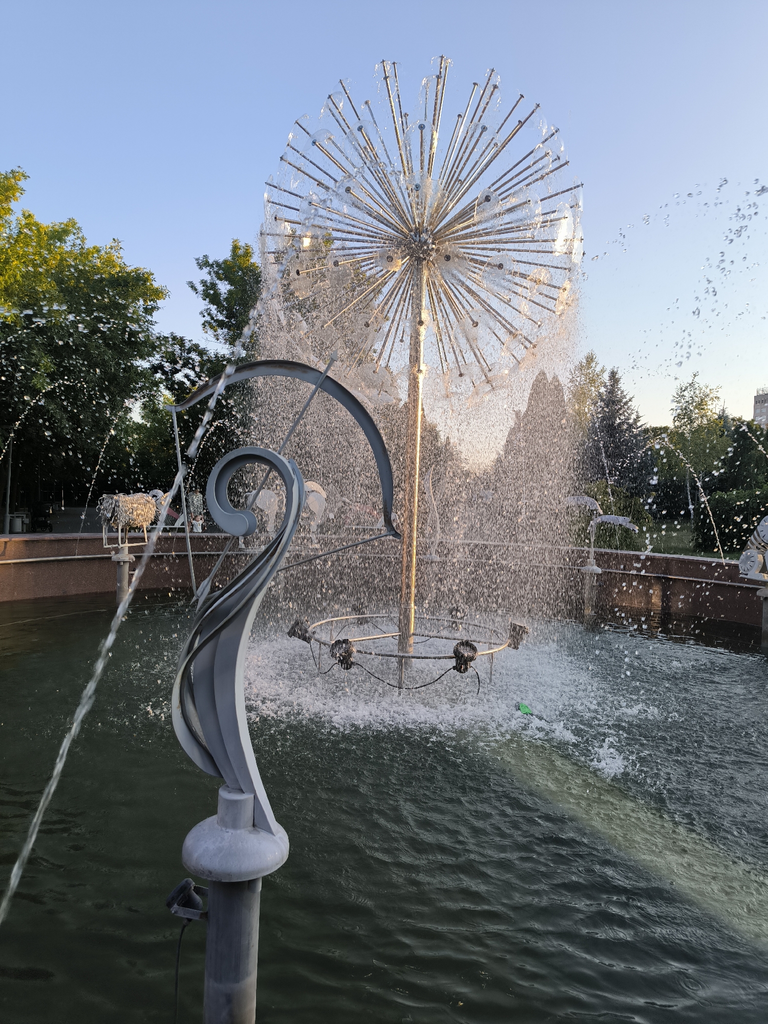The image shows a modern spherical fountain with water spraying outwards. The fountain has a metallic, dandelion-like structure at its center, with numerous thin rods radiating from a central point. Water jets stream from various points on this structure, creating a shimmering effect and a dense spray. In the foreground, a stylized, grey metallic sculpture with a swirling design is partially visible, emerging from the water. The water in the fountain basin is dark green and rippling, with several jets of water entering from the left side of the frame. In the background, lush green trees and a clear blue sky are visible. The scene suggests an outdoor public space, likely a park or plaza, during daylight hours. While not explicitly visible, this type of fountain is often found in urban settings. It's possible this is the fountain located in the central square of Chișinău, Moldova, a popular landmark known for its modern design and water features.