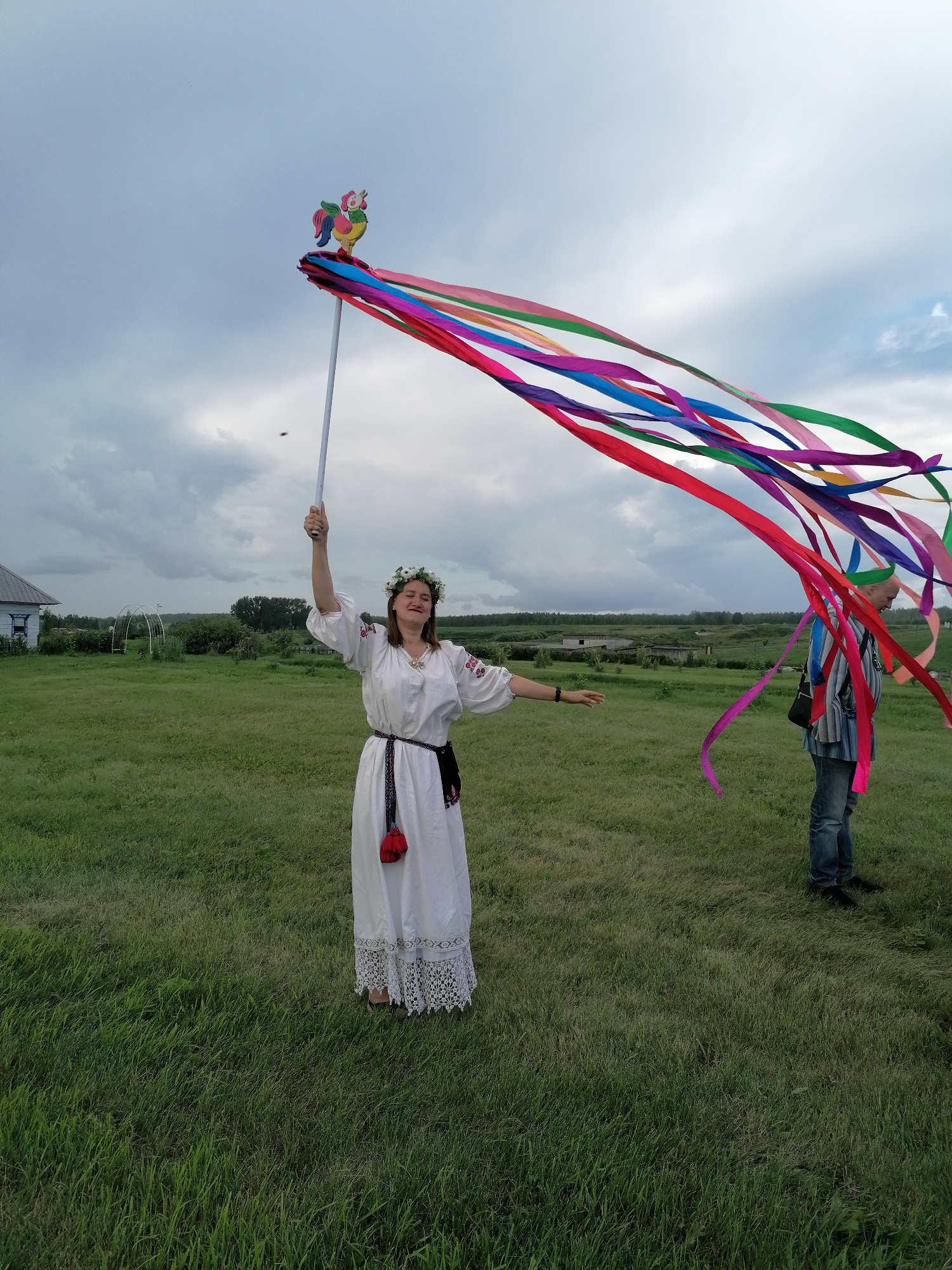 A woman in a white traditional dress with embroidery and a floral wreath on her head stands in a grassy field, holding a pole with colorful ribbons and a wooden rooster decoration. Her right arm is raised, and she appears to be moving the ribbons. A man in a striped shirt and jeans stands to her right, facing away from the camera. In the background, there is a white house with blue trim, some distant trees, and a cloudy sky. The scene is set in a rural environment, possibly in Mingali, Russia, given the presence of traditional attire and rural architecture.