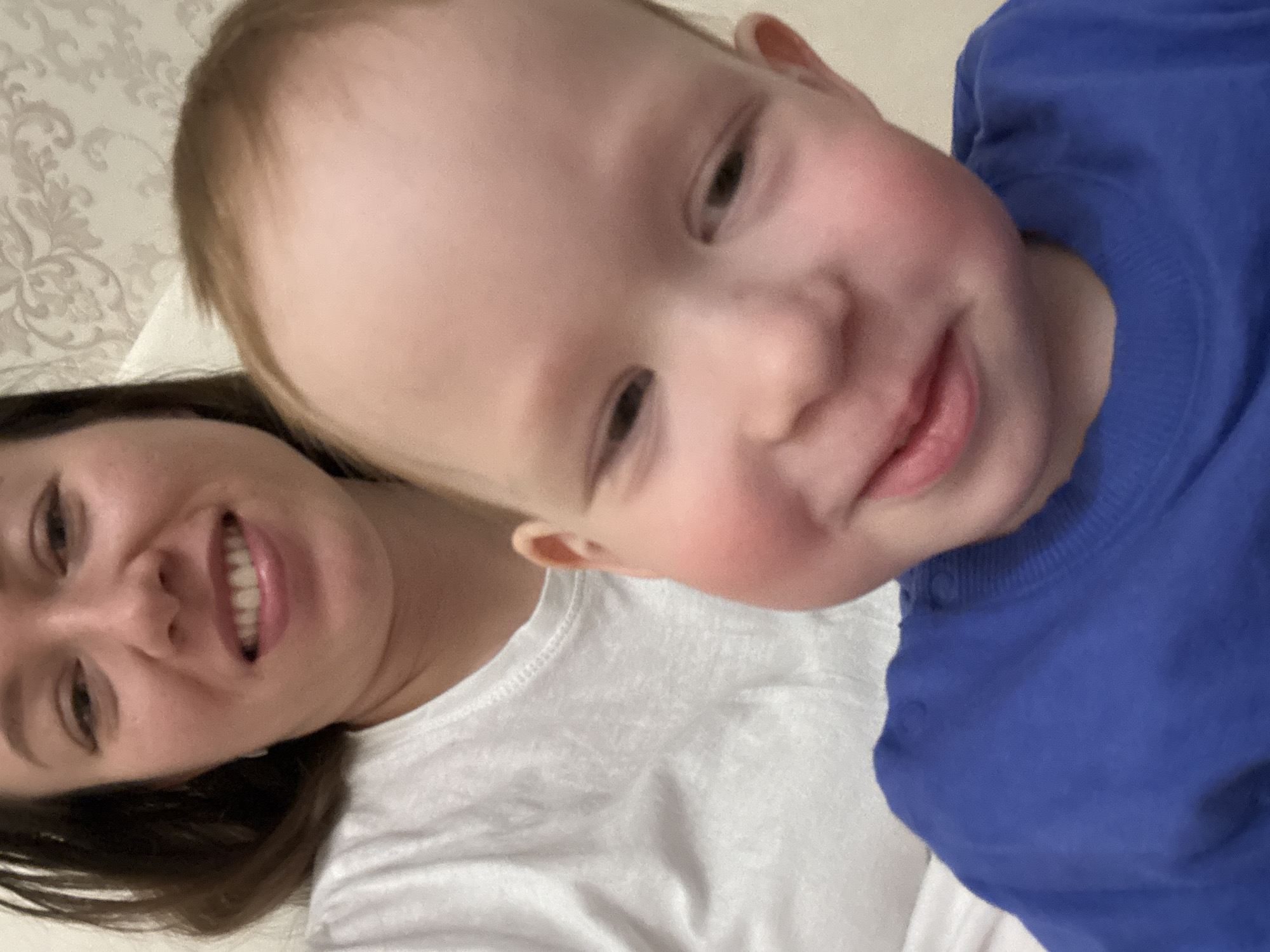 A selfie taken in a room with patterned wallpaper shows a woman and a baby. The woman is wearing a white t-shirt and has dark hair. The baby is wearing a blue shirt. They are close together and both smiling. The location is Ферма, Russia.