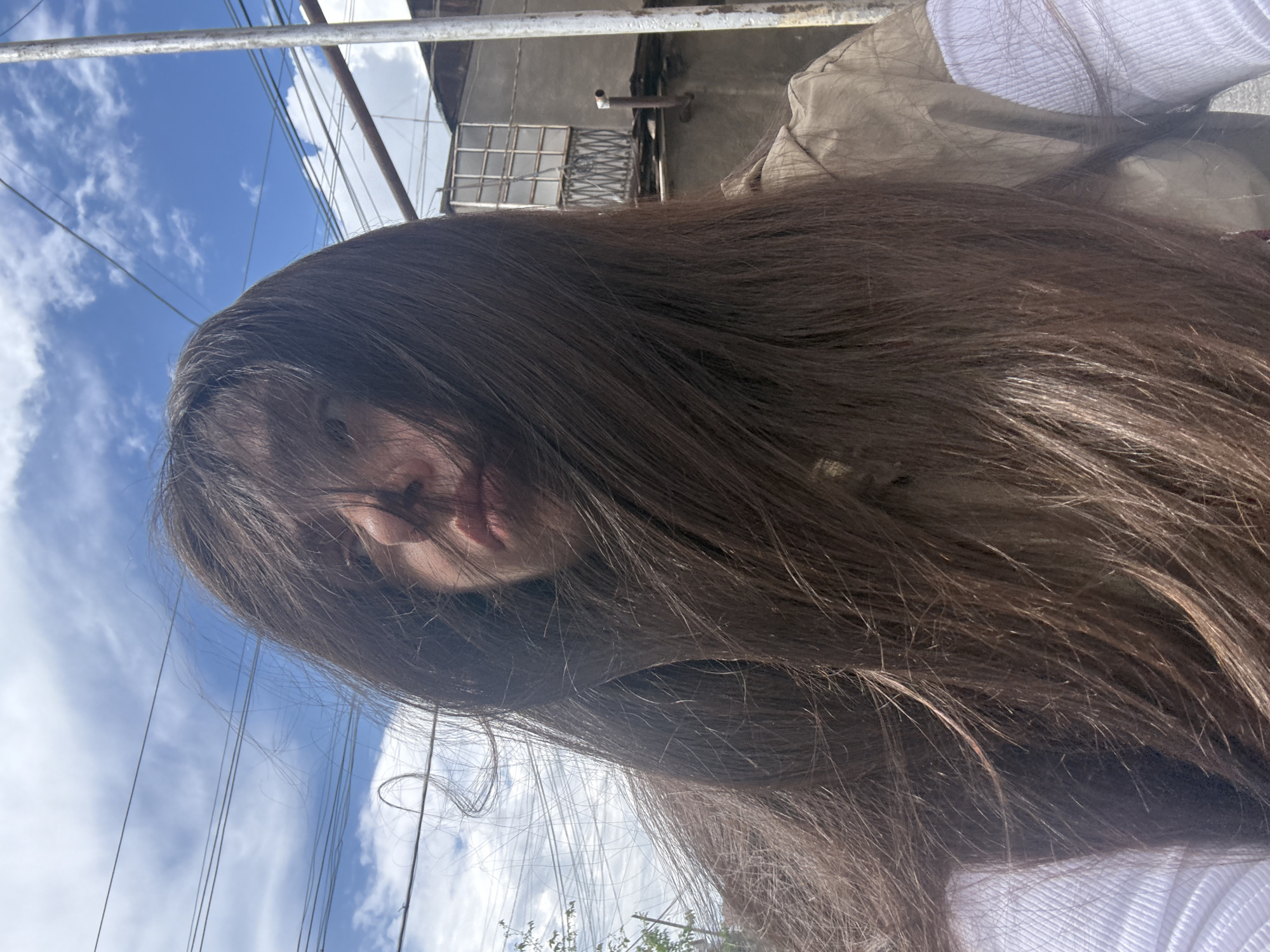 The photograph shows a person with long, dark brown hair obscuring their face, taken from a low angle looking upwards. The person is wearing a white shirt and a beige jacket. In the background, there is a blue sky with scattered white clouds, electrical wires, a building with a window, and a metal railing. The location is inferred to be Yerevan, Armenia.