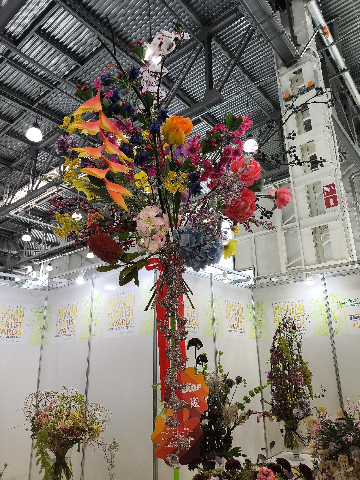 An overhead shot captures a vibrant floral arrangement suspended from a metal ceiling in an indoor exhibition hall. The flowers, a mix of artificial and possibly real, are predominantly orange, pink, yellow, and purple, with delicate white orchids and green foliage. The arrangement features cascading elements and is tied with a red ribbon and decorative metallic strings. Below the suspended display, several white exhibition booths are visible, adorned with banners promoting "Russian Florist Awards" and "Flowers Expo 2026." Various other floral arrangements and displays are situated on tables and stands within the booths. The environment appears to be a large-scale event space with industrial ceiling structures and lighting fixtures.